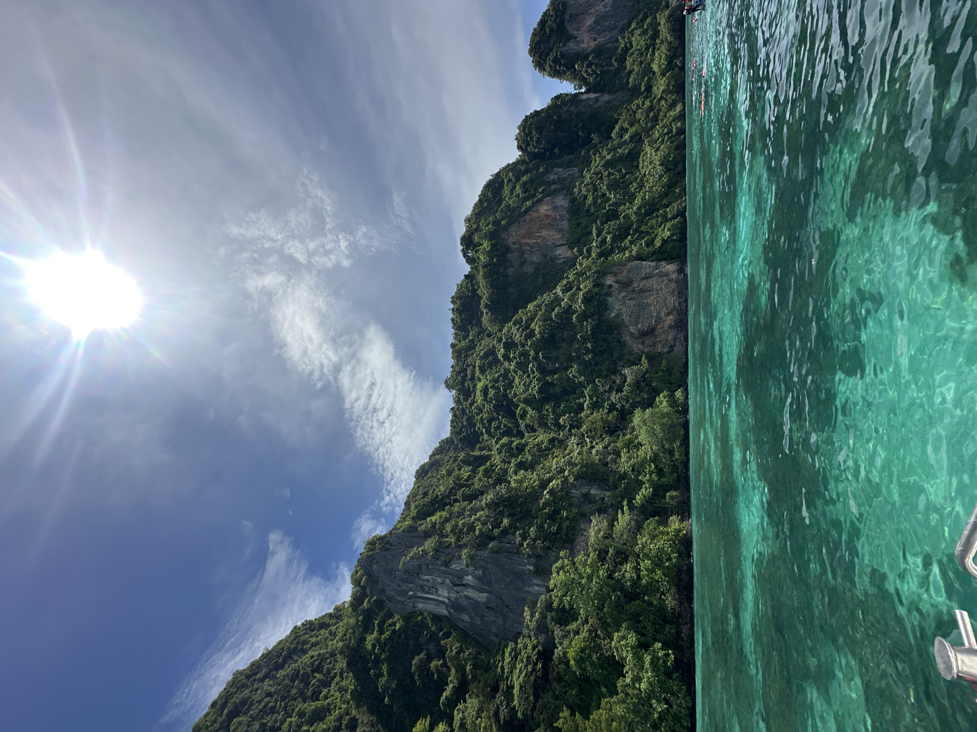 A high-angle, medium shot captures a vibrant tropical seascape with lush green cliffs dominating the background. The sun shines brightly from the upper right quadrant, casting a lens flare across a clear blue sky dotted with wispy white clouds. In the foreground, the turquoise water is exceptionally clear, revealing the sandy bottom beneath. The water's surface is rippled, with the sunlight creating a shimmering effect. Along the shoreline, the steep cliffs are covered in dense green foliage, with visible rock faces interspersed. In the distance, several small figures can be seen in the water, some wearing orange life vests, suggesting swimming or snorkeling activities. The left edge of the frame shows the metallic railing of a boat, indicating that the photo was taken from a vessel. The scene depicts a tranquil and idyllic tropical paradise.