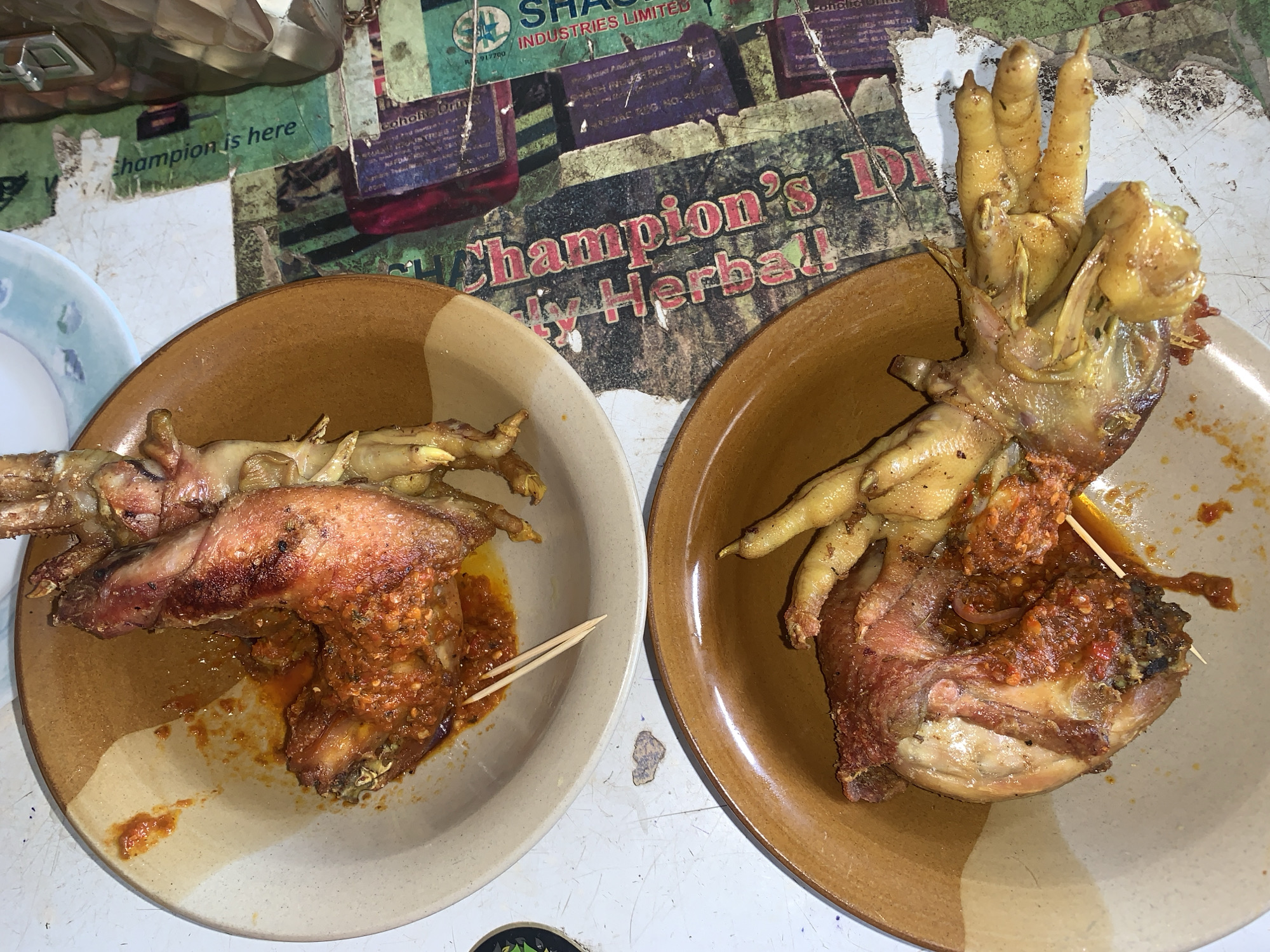 The image shows two plates of food, presumably taken in Jos, Nigeria, based on the background signage for "Champion's Herbal" and a local manufacturer, "SH Industries Limited" visible behind the plates. Each plate holds a piece of fried or grilled chicken covered in a red sauce and topped with what appears to be chicken feet. One plate has two toothpicks inserted into the food. The plates are brown and tan, and are placed on a surface with faded green and red lettering. There are also glimpses of what appears to be kitchenware in the upper left corner of the image.