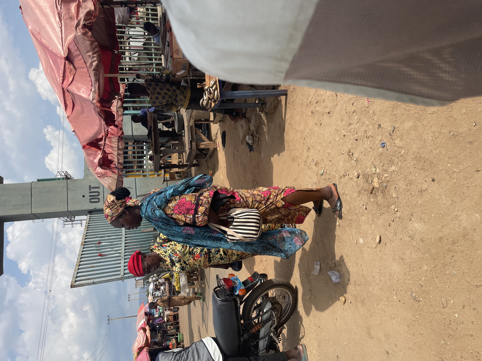 Two women, one wearing a red headscarf and the other a patterned headscarf, walk together along a dusty street in Akwanga, Nigeria. The woman in front carries a striped purse. They are near a gate with the word "OUT" on it. Street vendors with stalls under red awnings are visible in the background. A parked motorcycle stands in the foreground. The ground is brown dirt.