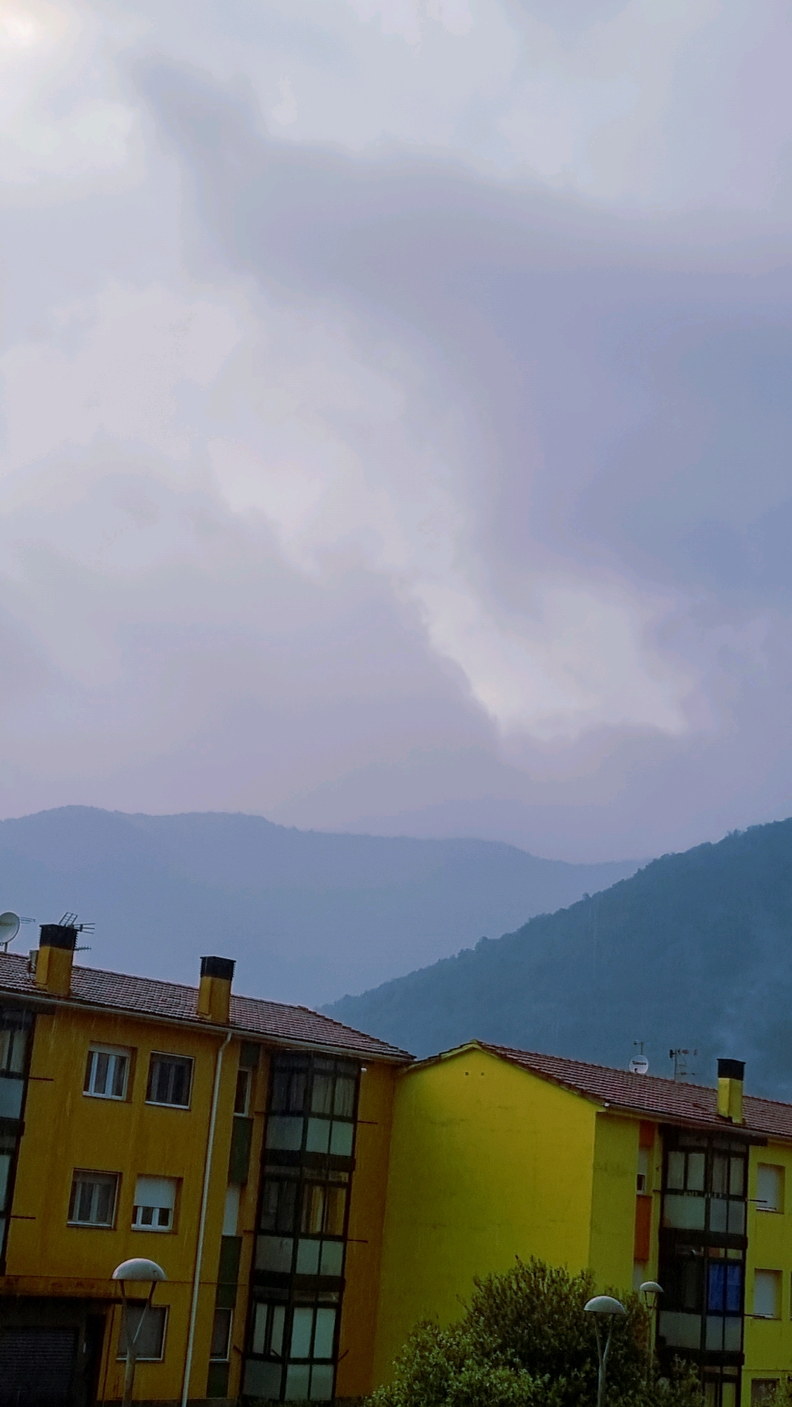 This is a medium-shot, vertical, exterior photograph taken in Sant Joan Les Fonts, Spain. The image captures a view of layered, hazy mountains in the background, partially obscured by clouds. In the foreground, a bright yellow, multi-story apartment building with red tiled roofs dominates the lower portion of the frame. The building has numerous windows, some with white shutters, and balconies with dark frames. Chimneys are visible on the roofline. Trees and a streetlamp are also present in front of the building. The sky above is overcast with soft, diffused light. The overall impression is a somewhat muted, possibly damp, atmosphere, suggesting overcast or rainy weather.