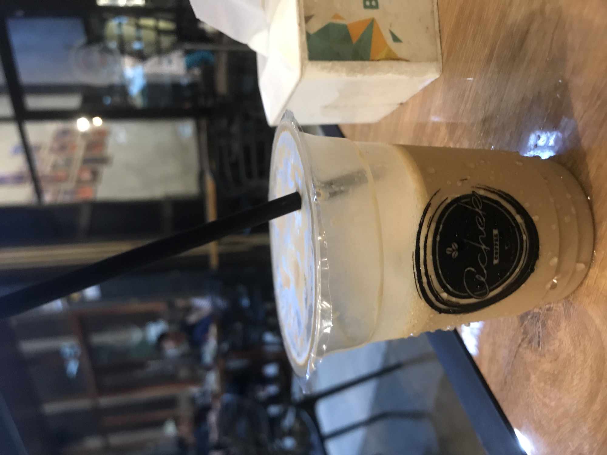 The image shows a close-up of an iced coffee beverage in a clear plastic cup with a black straw, sitting on a table. The cup has a black logo that reads "Cache Coffee". A tissue box is also visible in the background. The reflection in the glass table surface suggests an outdoor setting, possibly a cafe or restaurant in Lhokseumawe, Indonesia, with blurred figures of people in the background. The focus is on the beverage, and the scene is set in what appears to be a casual, public setting.
