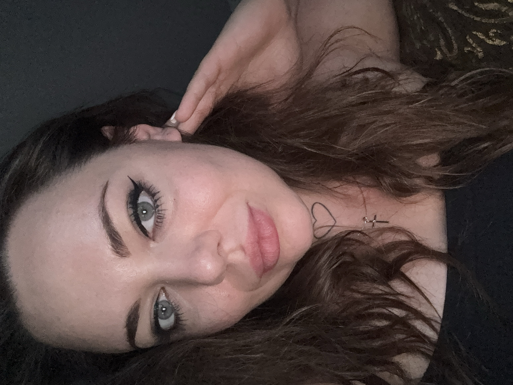 A woman is pictured in a close-up, head-and-shoulders shot, facing the camera. Her eyes are a light blue-gray and are adorned with dramatic winged eyeliner and mascara. She has full lips, tinted with a soft pink lipstick, and a subtle smile. Her hair is long, wavy, and dark brown. A small heart tattoo is visible on her neck, just below her jawline, and a delicate cross necklace rests on her collarbone. Her right hand is raised to her ear, with her fingers gently touching the lobe. She is wearing a black top. The background is dark and indistinct, providing no specific environmental details or context. There is no indication of the location being in Kremenchuk, Ukraine.