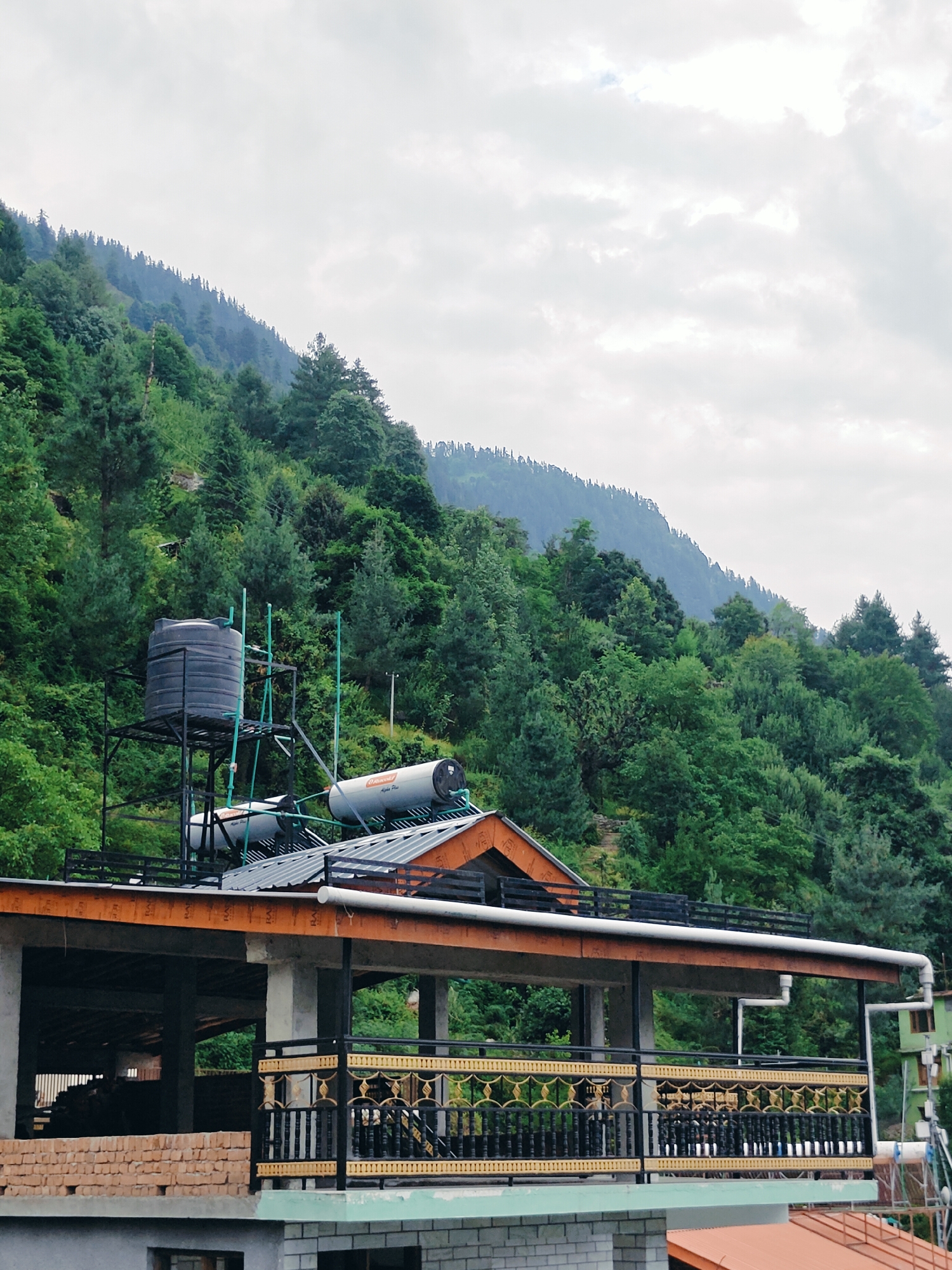 The image shows a building with a dark water tank on a metal structure, two solar water heaters on the roof, and a covered porch with a decorative railing. Behind the building, a lush green mountainside covered in dense trees rises towards a cloudy sky. The architecture suggests a location in a hilly or mountainous region, consistent with Vashisht, India, known for its natural beauty and hot springs. The building appears to be under construction or recently built, with exposed brickwork visible on the lower levels. The scene is static, with no people or animals present. The overall atmosphere is one of a serene, natural environment juxtaposed with functional, modern amenities.