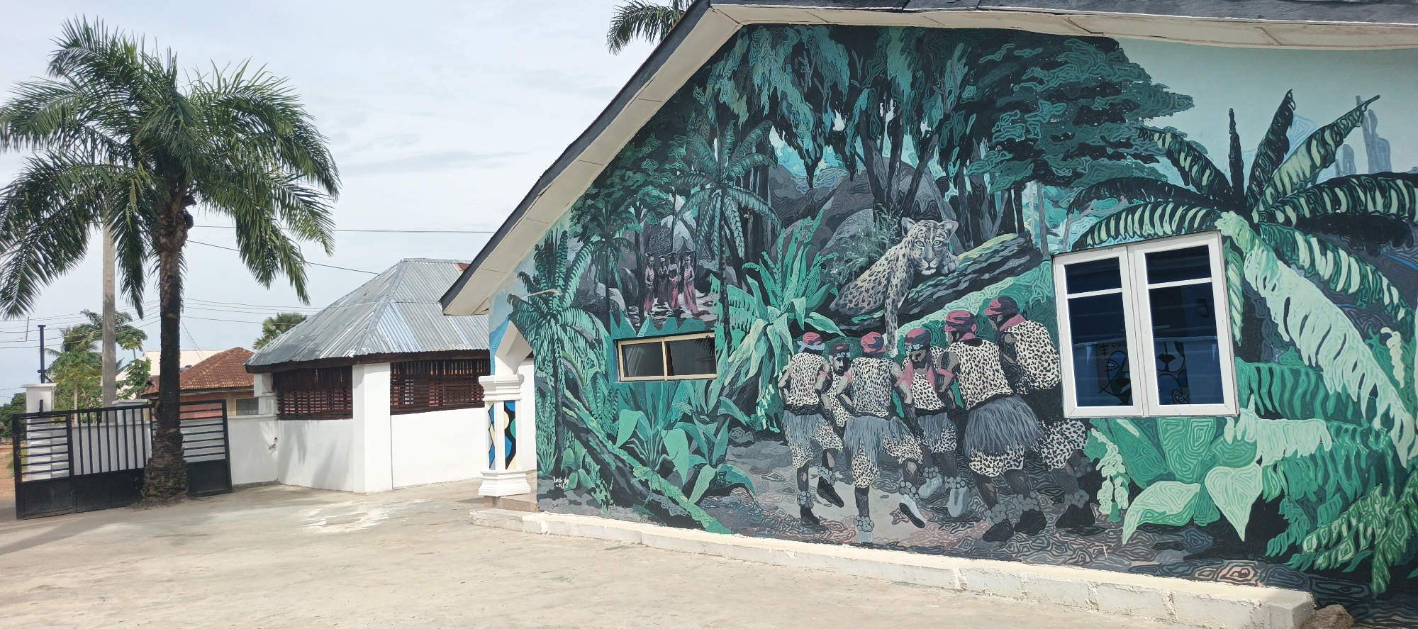 This outdoor image captures a vibrant building mural in Akwanga, Nigeria, under a bright, clear sky. The main subject is the side of a building completely covered by a large, colorful mural depicting a rich, dense jungle environment intertwined with cultural scenes.
The mural is dominated by various shades of green foliage, including numerous palm trees and large tropical leaves. Within this jungle setting, several figures are depicted. On the lower right, a group of approximately six to seven individuals, dressed in leopard-print tunics or tops, dark skirts, and red head coverings, appear to be engaged in a dance or ritualistic gathering. Above them and slightly to the right, a spotted big cat, resembling a leopard or jaguar, rests on a fallen log, looking towards the viewer. On the upper left section of the mural, a trio of women in traditional robes—pink, red, and light blue—and head coverings walk through the verdant landscape.
Two windows are integrated into the mural on the building wall: a smaller, rectangular window on the left, and a larger, double-paned window with blue-tinted glass on the right. At the building's corner, a decorative white column adorned with blue and black geometric patterns stands. Faint blue text, "Sunny Art 2023," is visible vertically on the side of this column, likely a signature and date.
In the background, to the left, another building with a corrugated metal roof and white walls is partially visible, featuring dark wooden lattice panels. A tall palm tree stands in the mid-ground, next to a black metal gate. The foreground consists of a wide, light-colored concrete or paved surface. Real palm trees are also seen against the light sky in the background, adding to the tropical atmosphere of the scene.