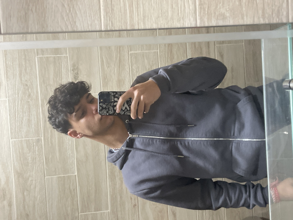 A male individual is depicted from the upper torso upwards, reflected in a mirror. He has dark, curly hair with a close-cropped fade on the sides. The subject wears a dark grey, full-zip hooded sweatshirt with a silver-toned zipper and a visible silver chain around his neck. His right hand holds a smartphone, encased in a black cover featuring a white bandana-style pattern, positioned to obscure the lower portion of his face (nose, mouth, and chin). A red and white braided bracelet is present on his left wrist. The environment reflected in the mirror consists of light beige, rectangular wall tiles arranged horizontally. A clear glass horizontal surface or shelf is visible in the foreground, with a thin vertical white strip (likely a mirror edge or frame) on the right side of the reflection. The scene is located in Cherni Vrah, Bulgaria.