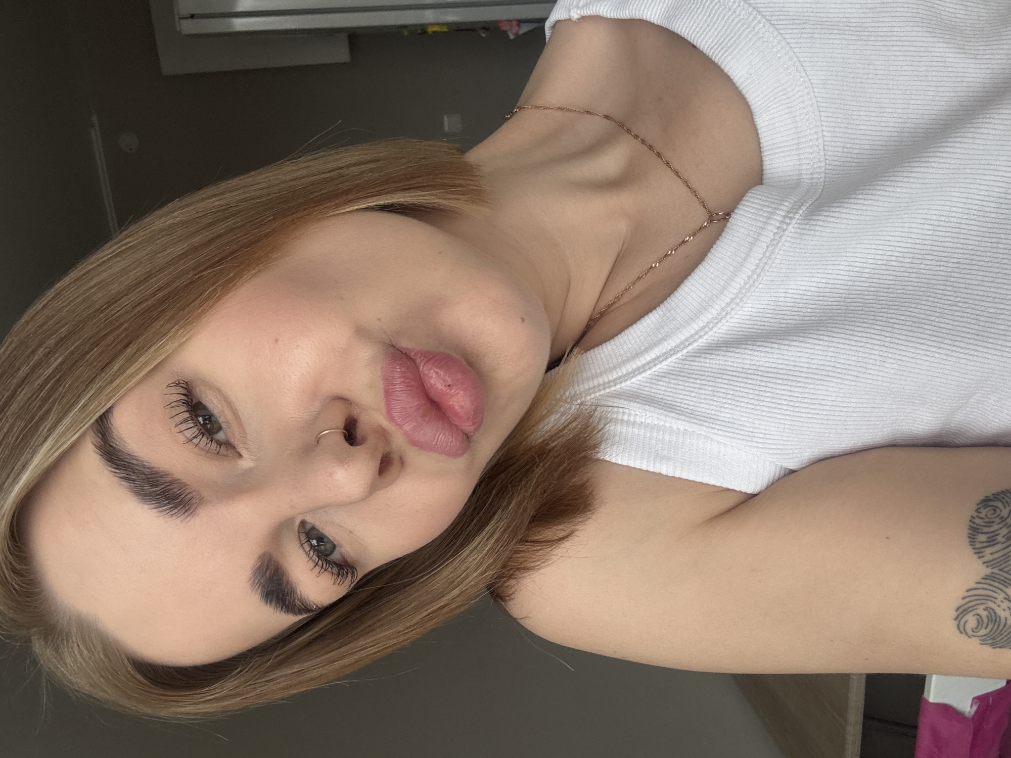 A close-up, rotated portrait of a woman with blonde bobbed hair. She has fair skin, defined eyebrows, long eyelashes, and is wearing a nose ring. Her lips are pursed in a pout. She is wearing a white ribbed tank top and a delicate gold necklace. A portion of a tattoo, appearing to be a fingerprint design, is visible on her arm. The background is out of focus, showing a grey wall and a glimpse of what appears to be a window fixture. There is no indication of location in the city of Chaikovsky, Russia.