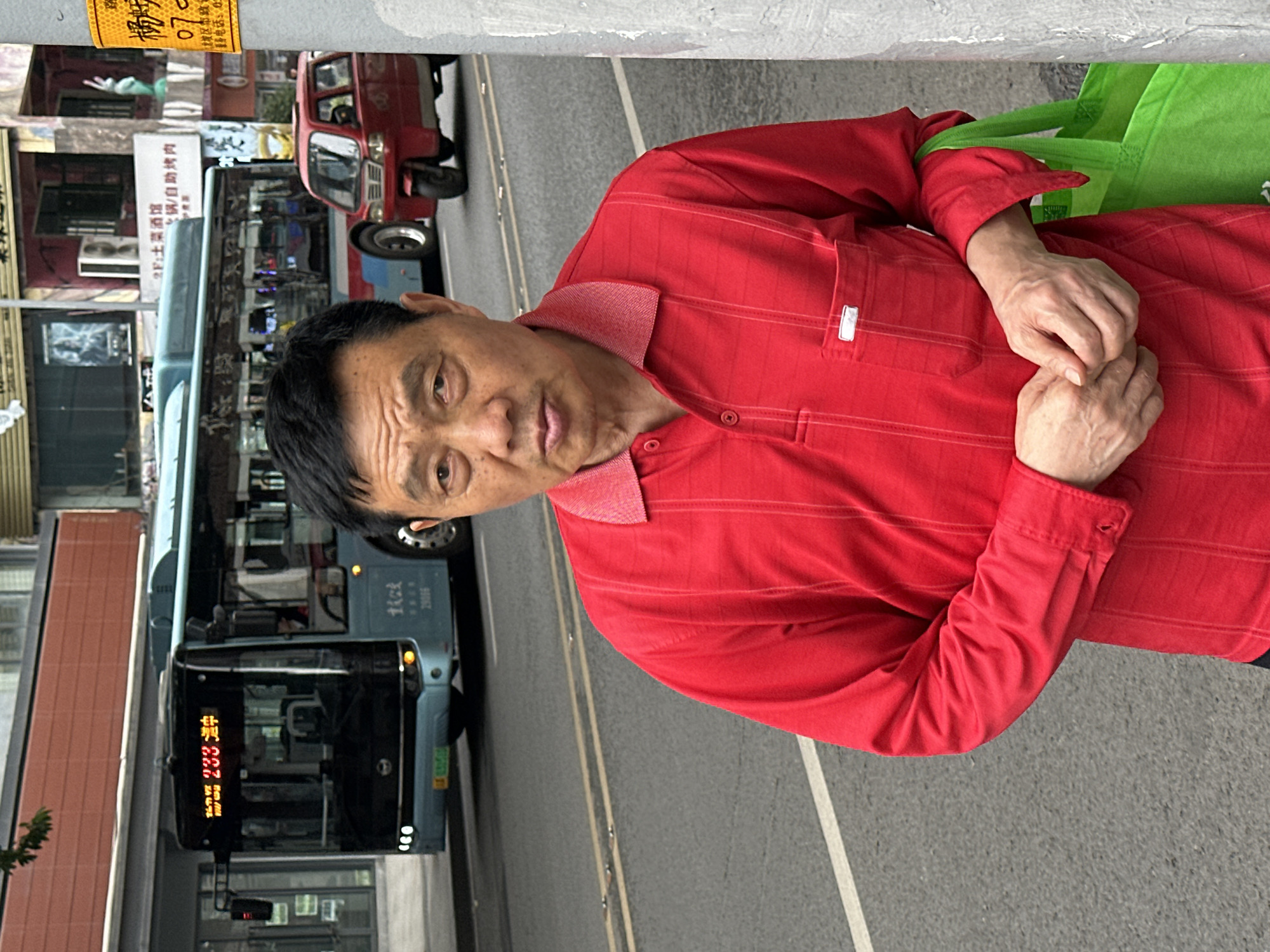 A man wearing a red shirt is standing on a city street next to a bus with the number 233 displayed on its front. The street is paved, and a sidewalk is visible on the left side of the frame. In the background, buildings with storefronts and a small red vehicle can be seen. Signs with Chinese characters are attached to the buildings. The bus has the text "29056" and "虾味" on its side. A sign with the text "2F:土菜酒馆 火锅/自助烤肉" is also visible. The location is in the city of Jiulongpo District, China.