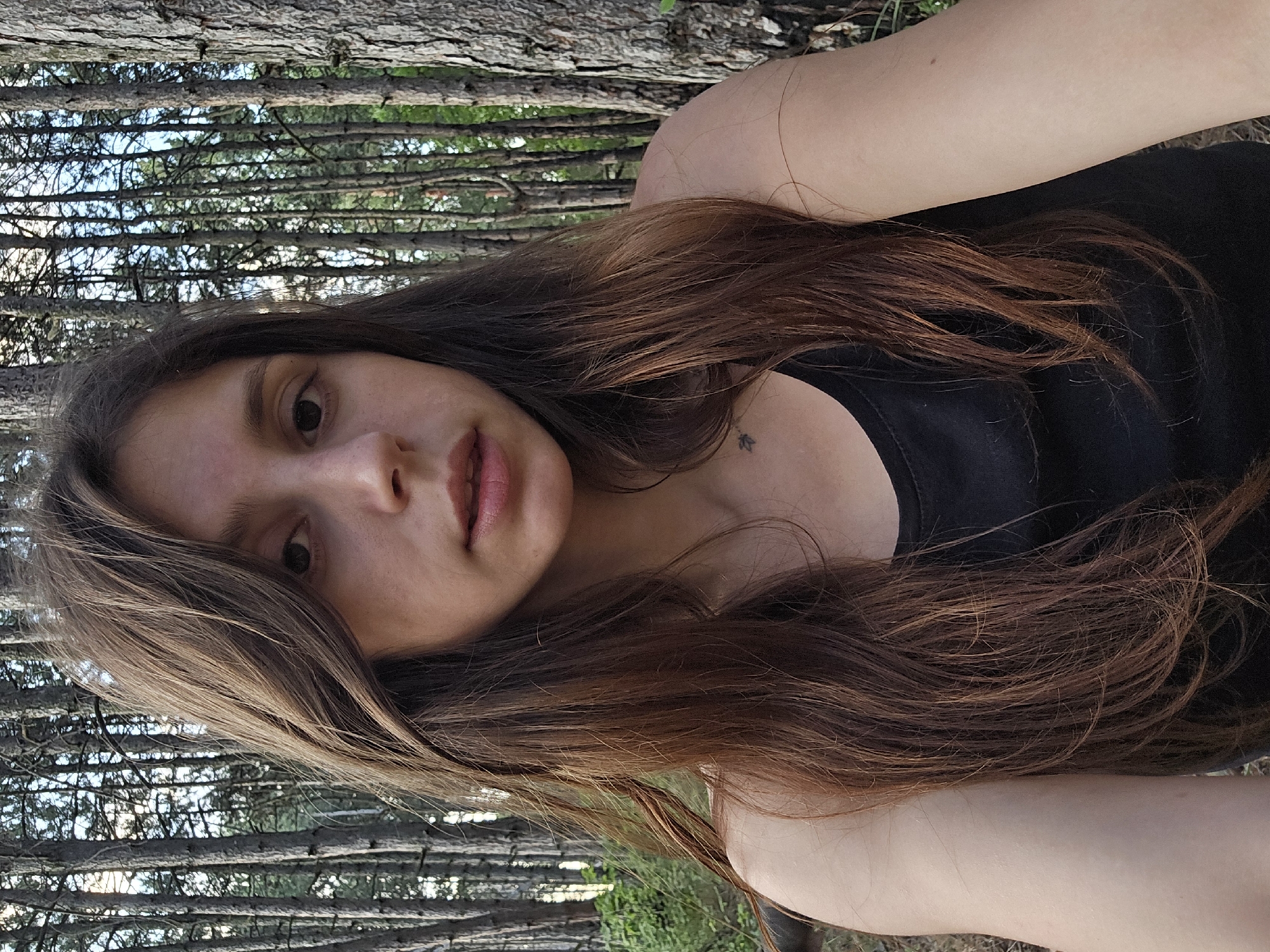 A selfie taken from the chest up shows a young woman with long, wavy brown hair standing in a forest. She is wearing a black tank top. The background consists of numerous tall, thin trees with rough bark and a sky visible through the branches. The lighting suggests it is daytime.