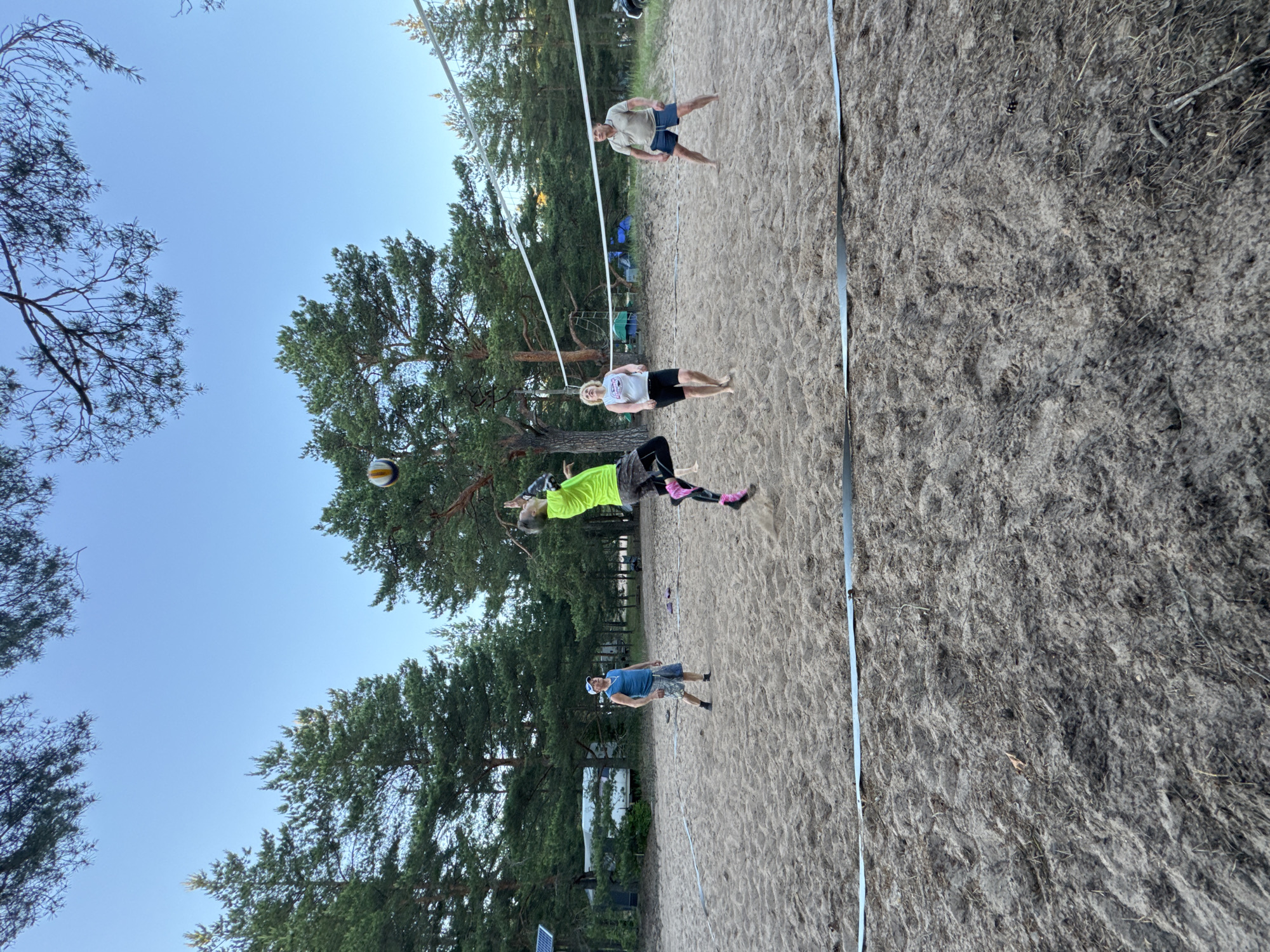 Four individuals are playing volleyball on a sandy court surrounded by pine trees. The court has a net stretched across it. One person in a neon green shirt is jumping to hit the ball. Another person in a blue tank top and shorts stands watching. A woman in a white tank top and black shorts and a man in a tan polo shirt and blue shorts are also on the court. In the background, there are tents and campers, suggesting this might be a campsite or recreational area. The sky is clear and blue. The scene appears to be in a natural outdoor setting with a focus on recreational activity.