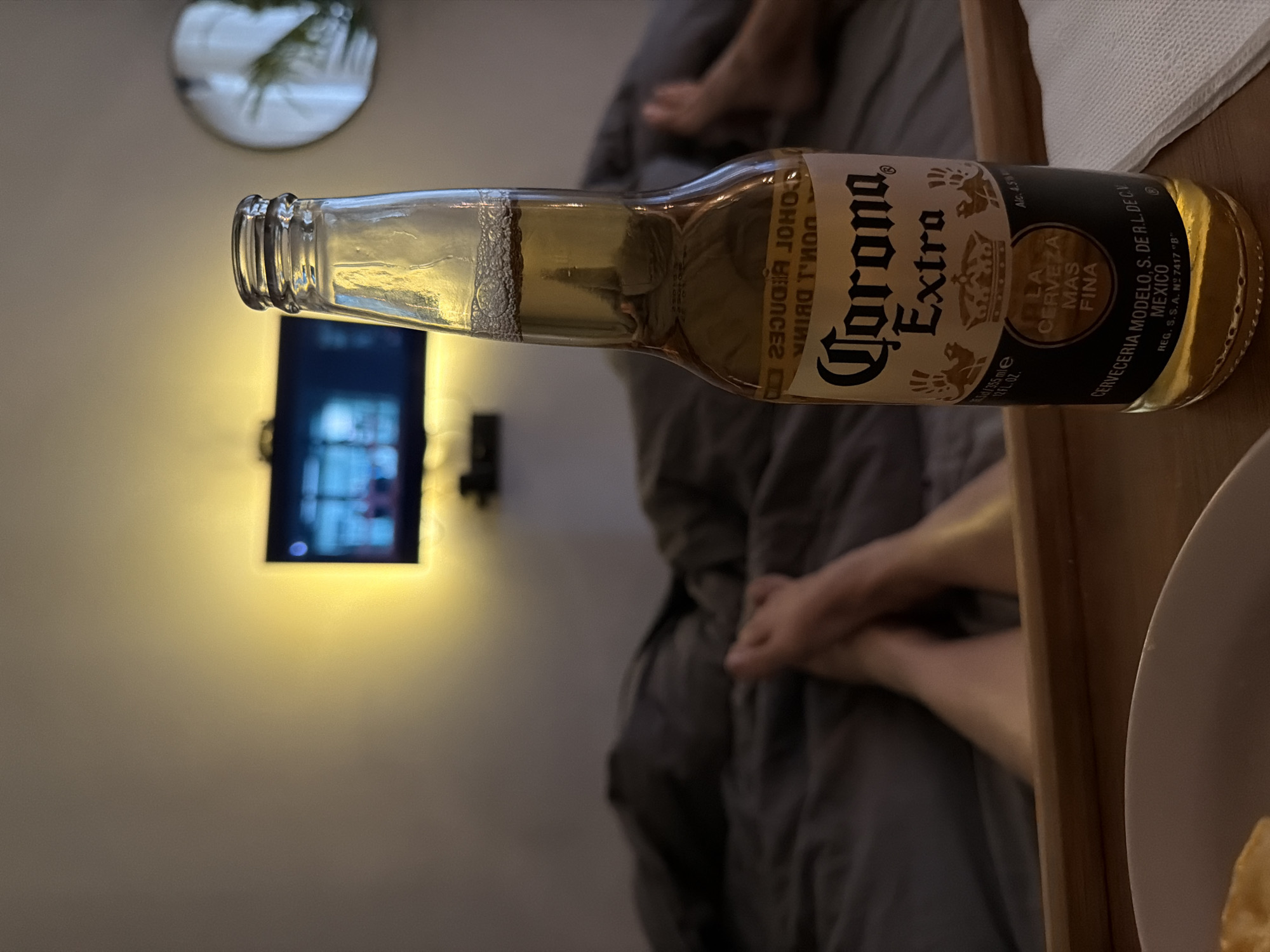 The scene is an indoor setting, likely a bedroom, as evidenced by the presence of a bed with grey bedding and a partially visible person lying on it. A bottle of Corona Extra beer sits on a wooden tray. In the background, a television is mounted on the wall. The lighting is warm and dim, creating a relaxed ambiance. A round, decorative object hangs on the wall to the left of the television. The location is unspecified, and it is not possible to determine if the image was taken in Khimki, Russia.