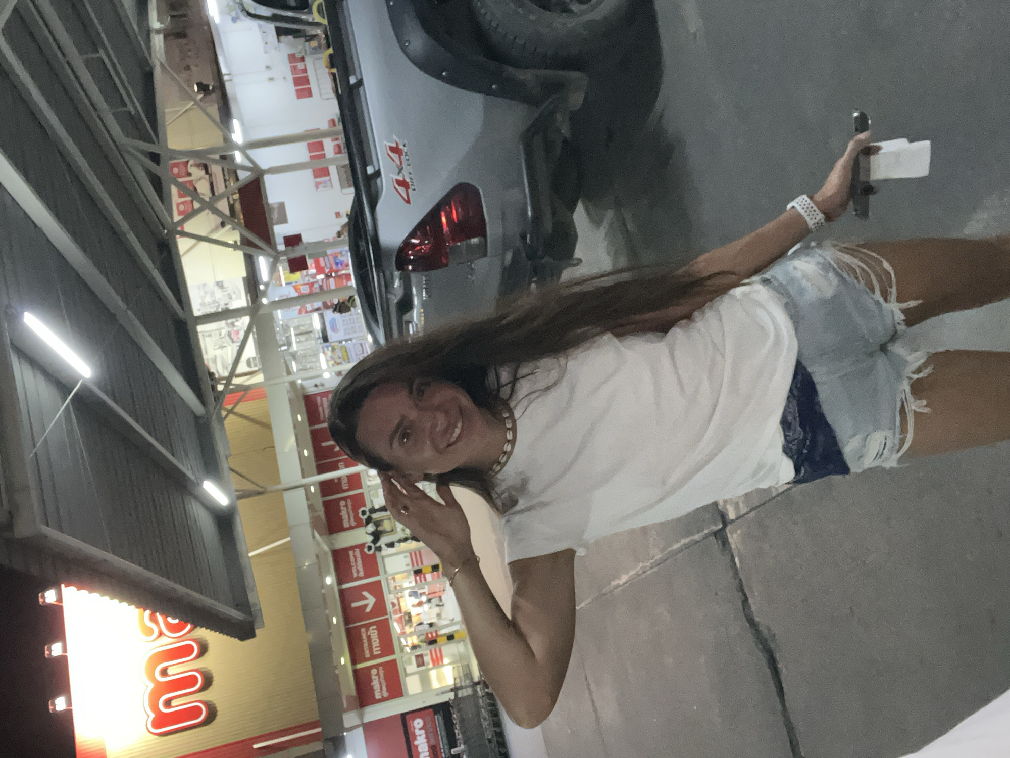 A woman is standing in front of a Makro store in Baan Bo Phut, Thailand. The woman is wearing a white t-shirt, denim shorts, and a white watch. She is holding a receipt in her right hand. Behind her, there is a silver pickup truck with "4x4 OFF ROAD" written on the tailgate. The Makro store has a large red and white sign with the store's logo. The image appears to be taken at dusk, as the lighting is dim and artificial lights are on.
