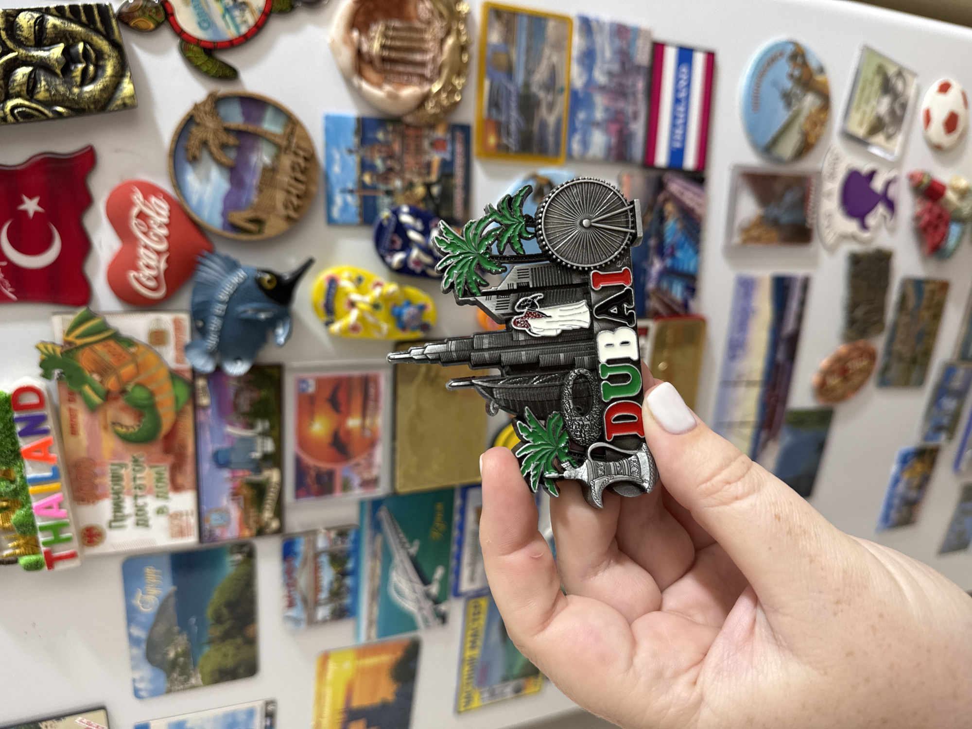 A person's hand is holding a souvenir magnet shaped like the Dubai skyline. The magnet features miniature replicas of the Burj Khalifa, a Ferris wheel, palm trees, and a traditional Arabic coffee pot, with the word "DUBAI" emblazoned in colorful letters. The background of the image is a white refrigerator door covered with various other souvenir magnets, including one depicting the Turkish flag, a golden Egyptian mask, a heart-shaped Coca-Cola logo, and multiple travel-themed magnets from different locations. The overall scene suggests a collection of travel memorabilia. The location is identified as Mikhaylovsk, Russia, based on the provided information.