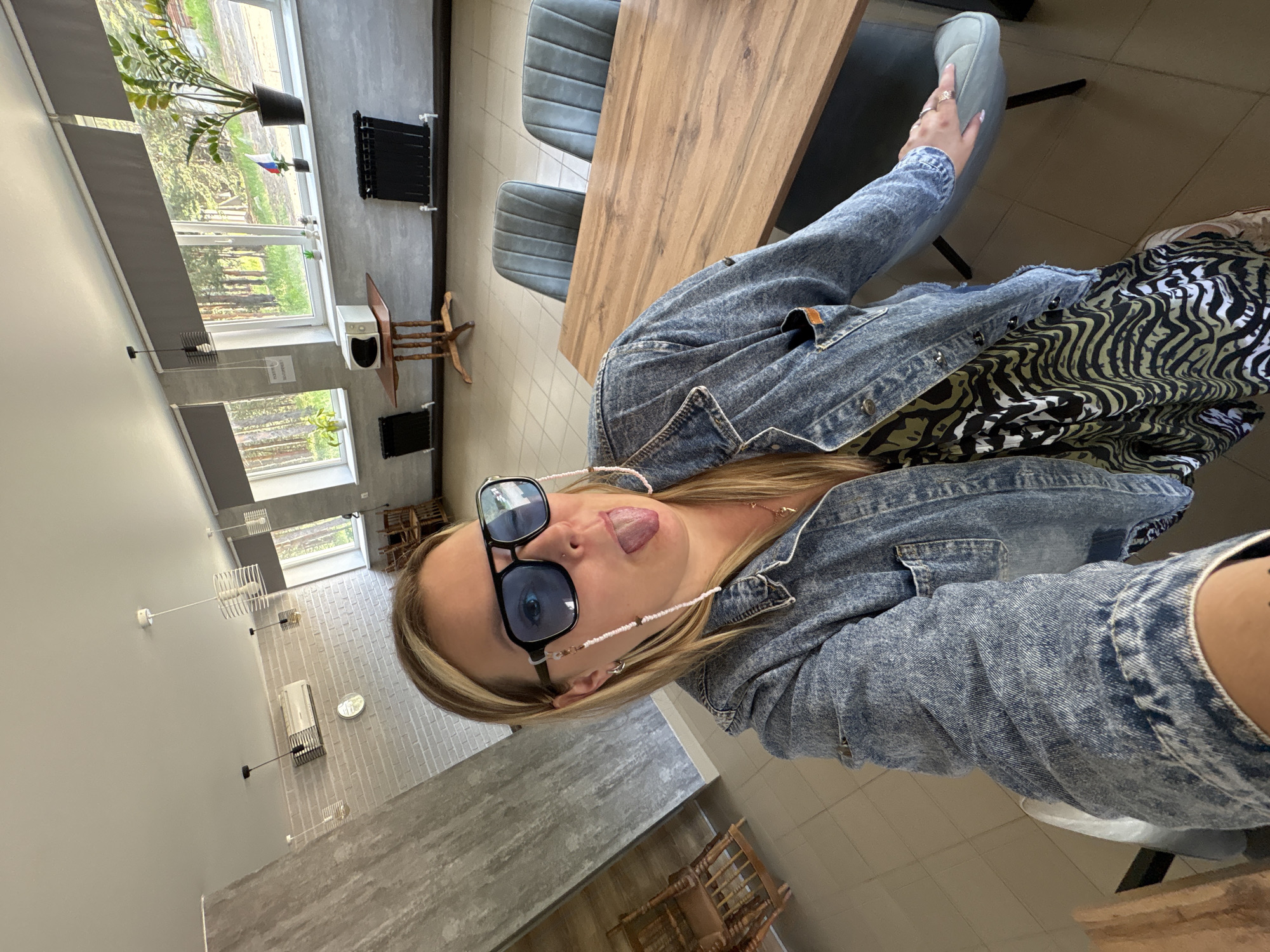 A woman with blonde hair is sitting at a wooden table in a room with large windows. She is wearing sunglasses and a denim jacket over a dress with a zebra-like print. Her tongue is sticking out. The room appears to be a dining area or common space, with chairs around the table, radiators along the wall, and a microwave on a small table. Outside the windows, trees and foliage are visible, suggesting a location surrounded by nature, possibly in or near Noviy, Russia.