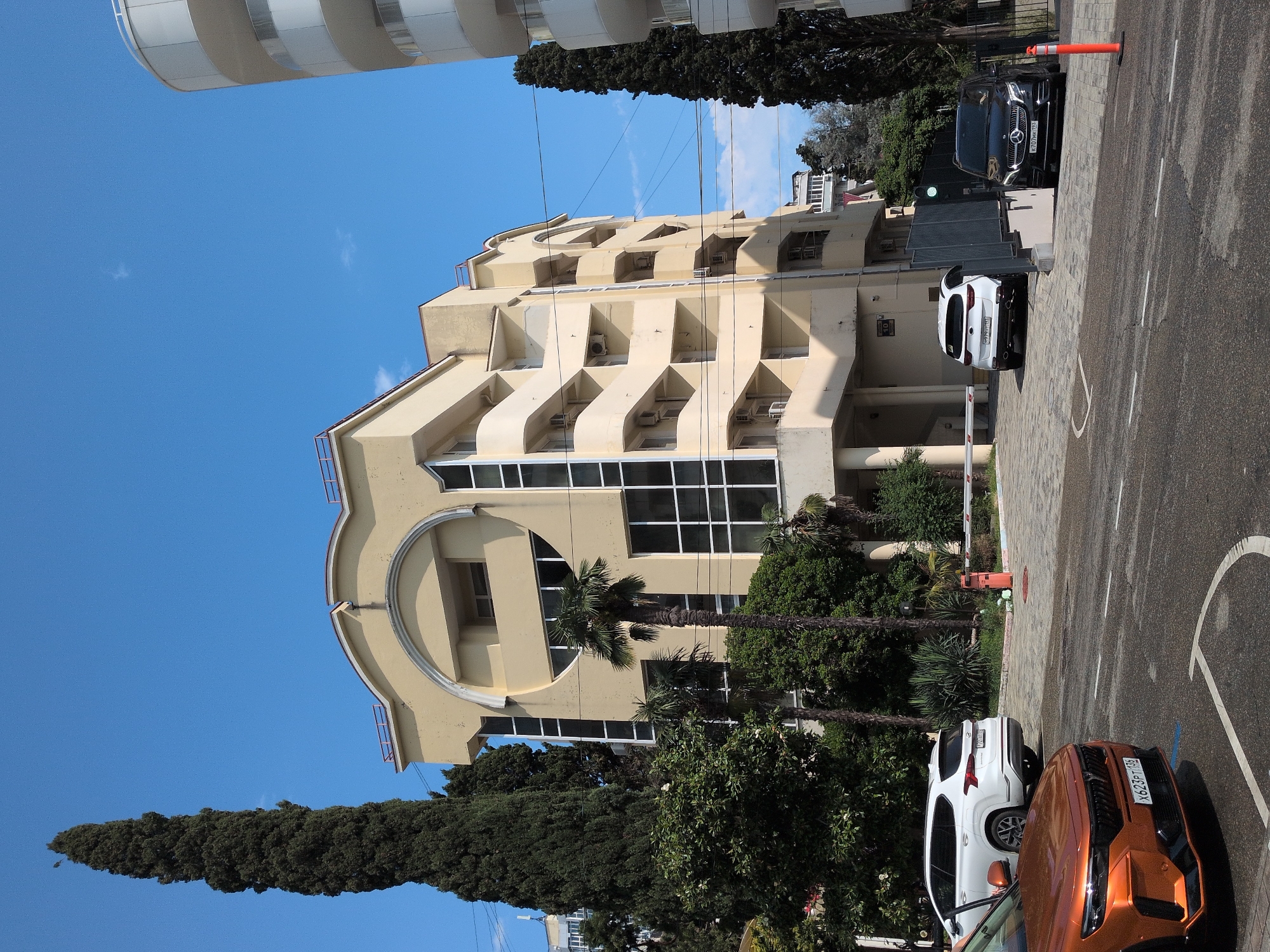 The image shows a street scene in Sochi, Russia, under a clear blue sky, suggesting it is daytime with fair weather. The main subject is a light-yellow, multi-story building with a distinctive rounded architectural feature on its facade. Balconies with railings are visible on several floors, as are air conditioning units. Palm trees and other lush green trees surround the building and are also visible in the foreground.
In the foreground, there are several cars parked on an asphalt surface marked with white lines. Two cars are particularly prominent: a white SUV and an orange car. A black Mercedes-Benz is also visible parked further back. The cars appear to be in a parking lot or a street with designated parking areas.
A red and white barrier gate is positioned across a paved area, suggesting a controlled entry or exit point. An orange traffic cone is placed near the barrier gate. The scene appears to be calm and uneventful, with no people visible. The overall impression is of a well-maintained urban or resort area.
License plates are partially visible on the cars, but the text is not clearly legible to be transcribed.