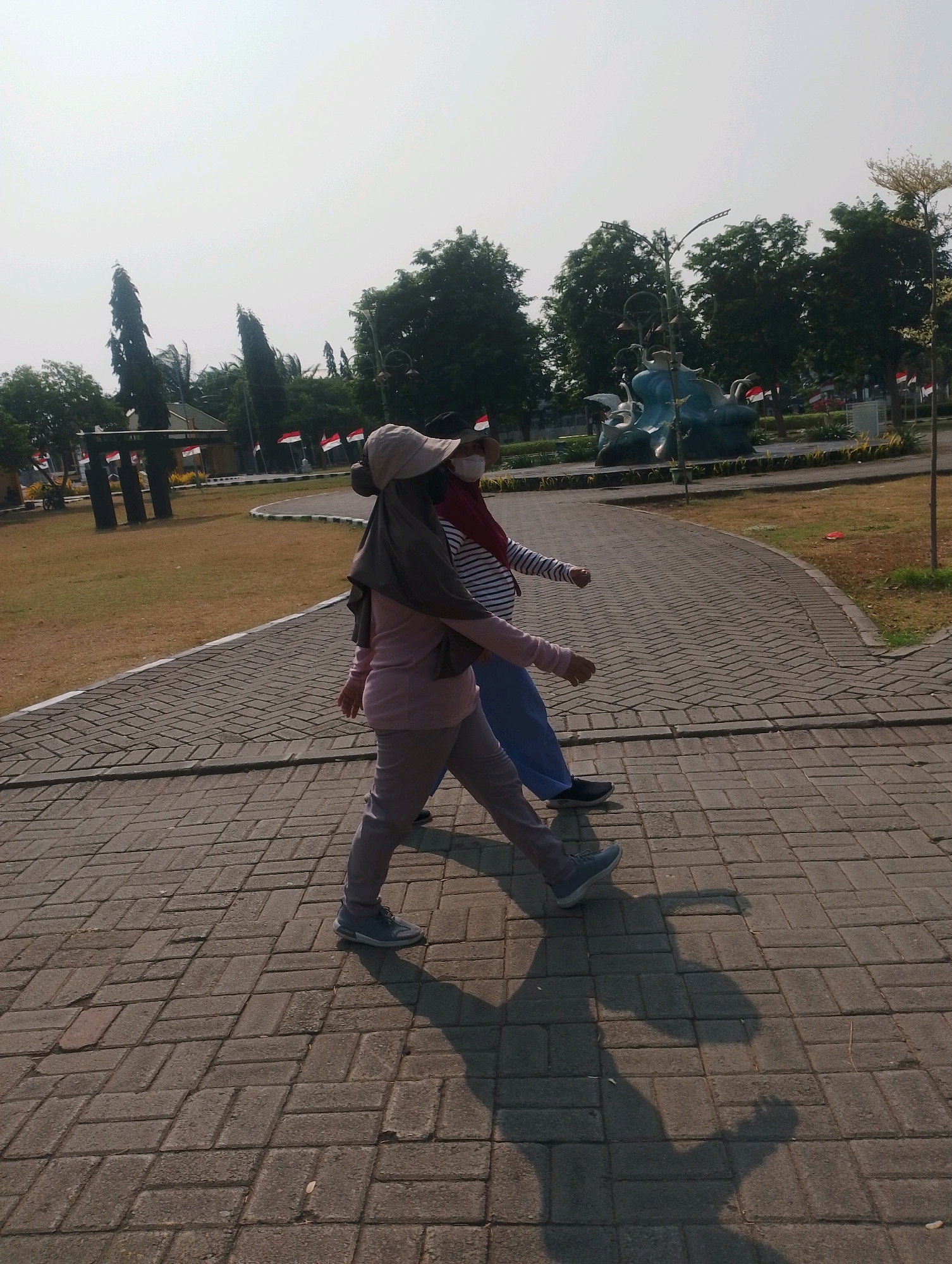 Two individuals are walking along a paved pathway in what appears to be a park or public garden. The person in the foreground, wearing a light-colored hat, a grey hijab, a pink long-sleeved shirt, and grey pants, is walking towards the right side of the frame. Behind them, another individual wearing a striped shirt and blue pants is also walking in the same direction. Both individuals have their faces obscured by masks. The pathway is constructed of interlocking grey bricks laid in a herringbone pattern. The environment features a grassy area to the left of the pathway, and in the background, there are trees, some with Indonesian flags displayed, and a sculpture of birds. The sun casts distinct shadows of the individuals onto the brick pathway. The scene suggests a daytime activity in an outdoor public space in Probolinggo, Indonesia.