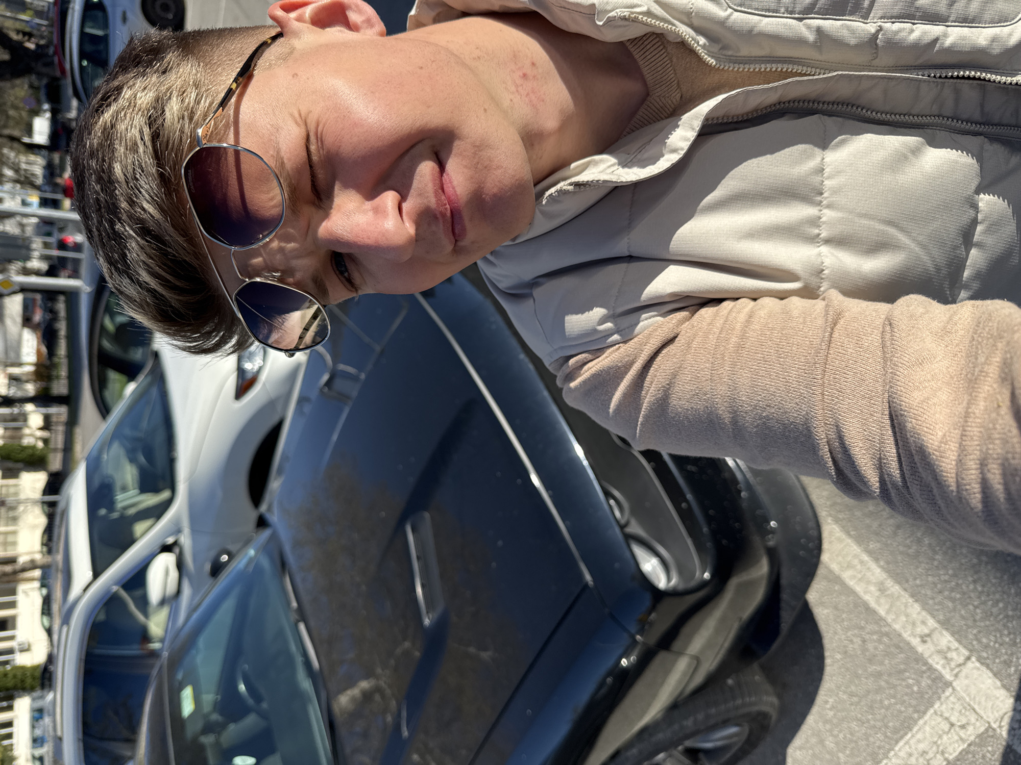A selfie-style photograph shows a person with short, light brown hair and sunglasses, wearing a light beige vest over a similar-colored sweater. The person is smiling slightly and appears to be standing next to a black car. Parked beside the black car is a white vehicle. The scene suggests an urban environment, possibly in Plovdiv, Bulgaria, given the location request. The background is somewhat blurred but suggests a street with buildings and possibly other parked vehicles.