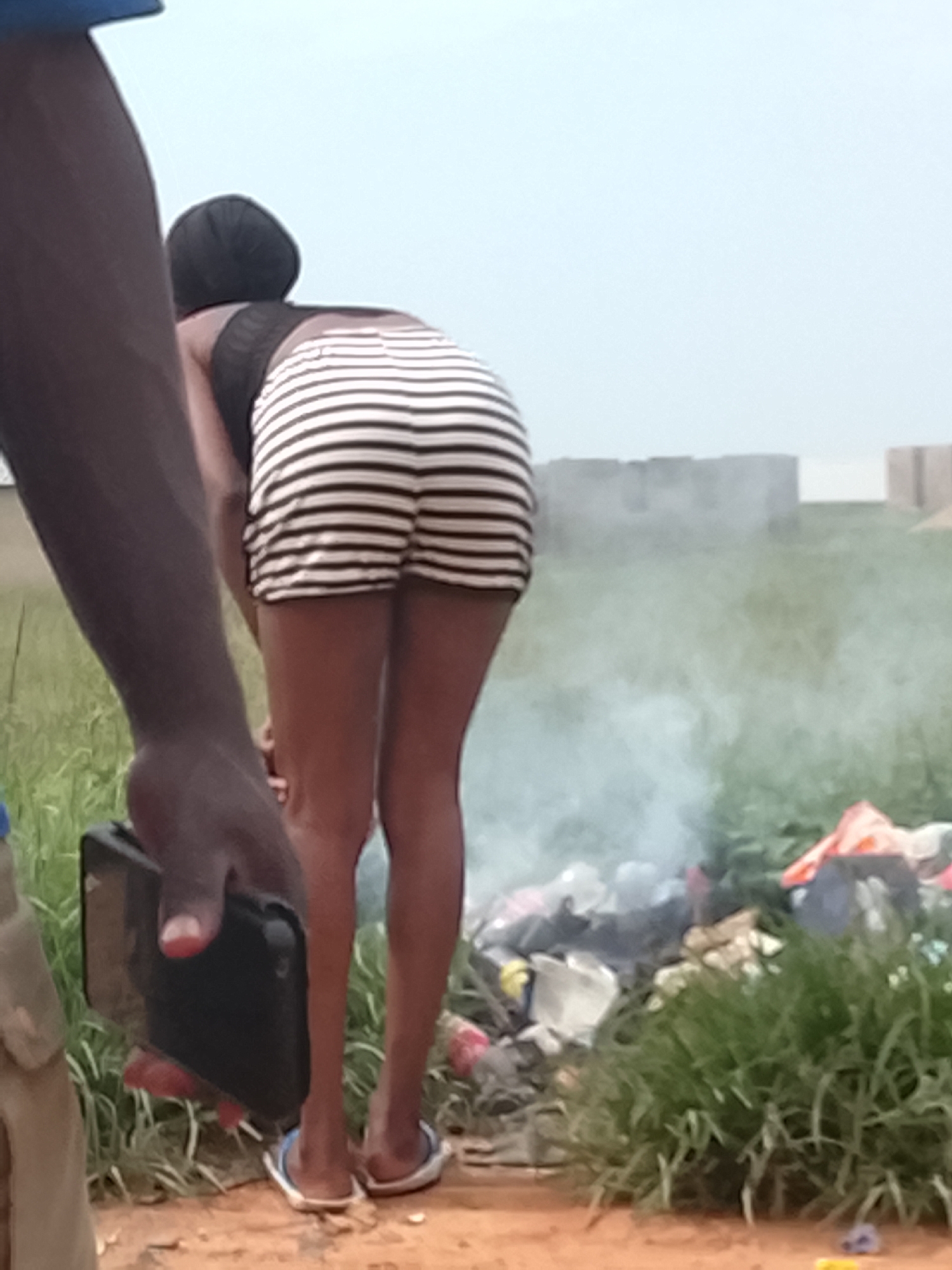 A person, viewed from the back, is bent over near a pile of burning trash. The person is wearing a black and white striped skirt and flip-flops. Another person's arm, holding a black phone, is visible in the foreground. The environment appears to be outdoors with dry grass and dirt. The sky is overcast and light blue. In the background, there are some unfinished brick structures. Smoke from the fire is visible.