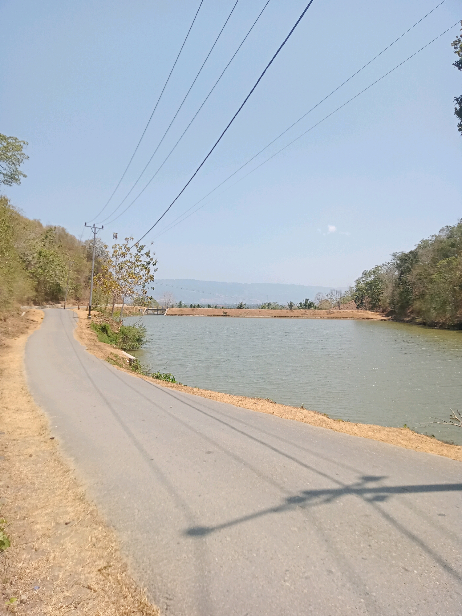 A paved road curves along the edge of a body of water, bordered by dry, grassy hills and trees. Overhead, power lines stretch across the clear blue sky. In the distance, mountains are visible under the haze. The scene appears to be a rural area, likely in Luhu, Indonesia, given the style of the landscape and infrastructure. There are no people or animals visible in the image. The overall impression is one of a quiet, sun-drenched landscape.
