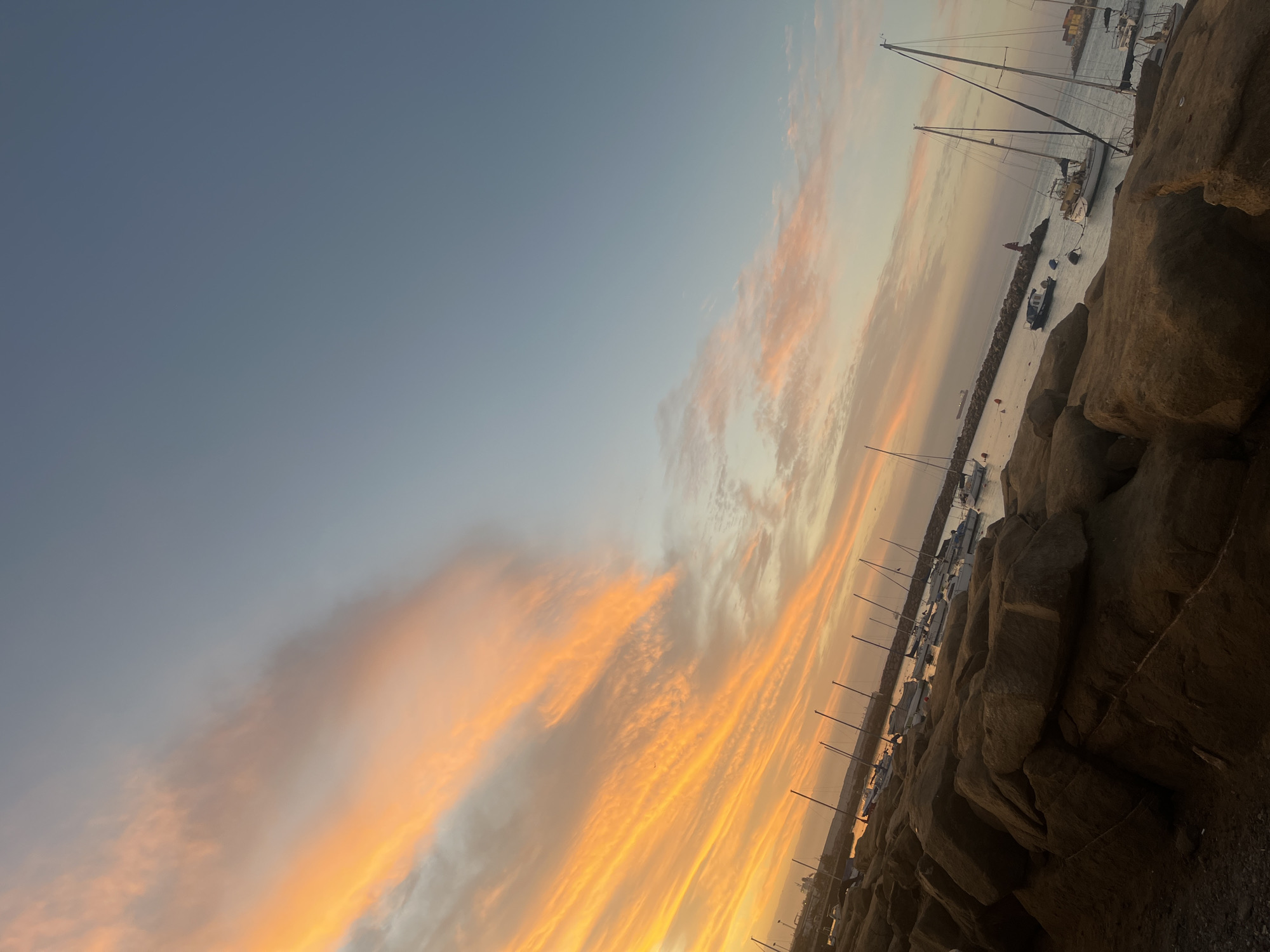 The image shows a coastal scene in Arica, Chile, likely at dusk or dawn, with a vibrant orange and yellow sky partially obscured by gray-blue clouds. To the right of the frame are large, dark brown rocks that appear to be part of a breakwater or shoreline. Beyond the rocks is a body of water, presumably the Pacific Ocean, where numerous sailboats and smaller boats are docked. The masts of the sailboats are visible against the colorful sky. There's also a small tower of a lighthouse. No people are visible in the image.