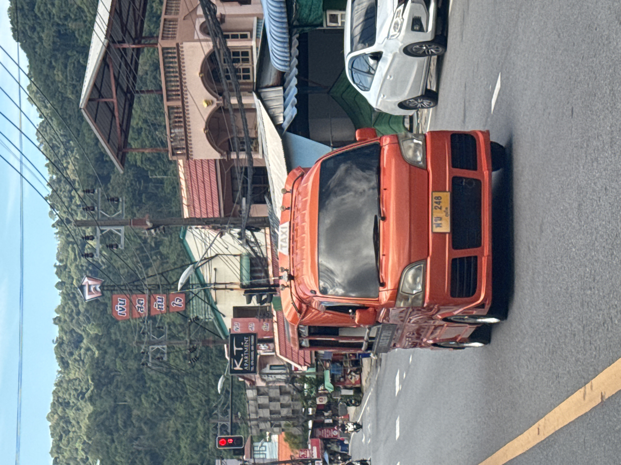 An orange, three-wheeled taxi with a "TAXI" sign on its roof drives down a paved road. The taxi has a yellow license plate with Thai characters and the number "248". A white sedan is to the right of the taxi. In the background, there are buildings with balconies and roofs, a lush green hillside with trees, and a blue sky with some clouds. There are power lines crisscrossing the scene. Traffic lights and signs are visible on the left side of the road.