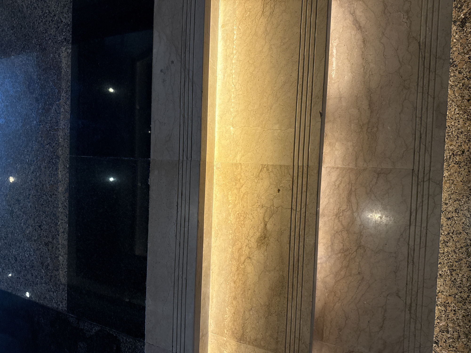 A close-up, slightly angled view shows a series of adjacent stone surfaces, possibly cladding or flooring, in Nusa Dua, Indonesia. The left side features a dark, reflective stone, interspersed with a speckled, coarse stone. Moving right, a lighter, beige-colored stone with subtle marbling is seen, followed by a pale gray marbled stone. The rightmost surface consists of small speckled gravel-like aggregates in a mix of light and dark colors. Vertical grooves accentuate the transitions between the stones. The lighting appears artificial, casting reflections on the polished surfaces. The overall scene suggests an interior or architectural detail.