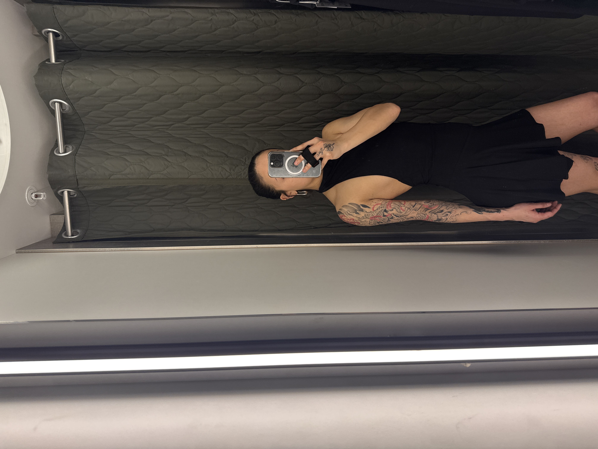 A photograph taken within a dressing room or fitting room setting. A person is holding a cell phone up to their face to take a selfie, wearing a black dress and exhibiting tattoos on their left arm. The background shows a quilted curtain and the overall environment suggests a retail space with dressing room facilities. The location in the city of London, United Kingdom, cannot be determined from the given image.
