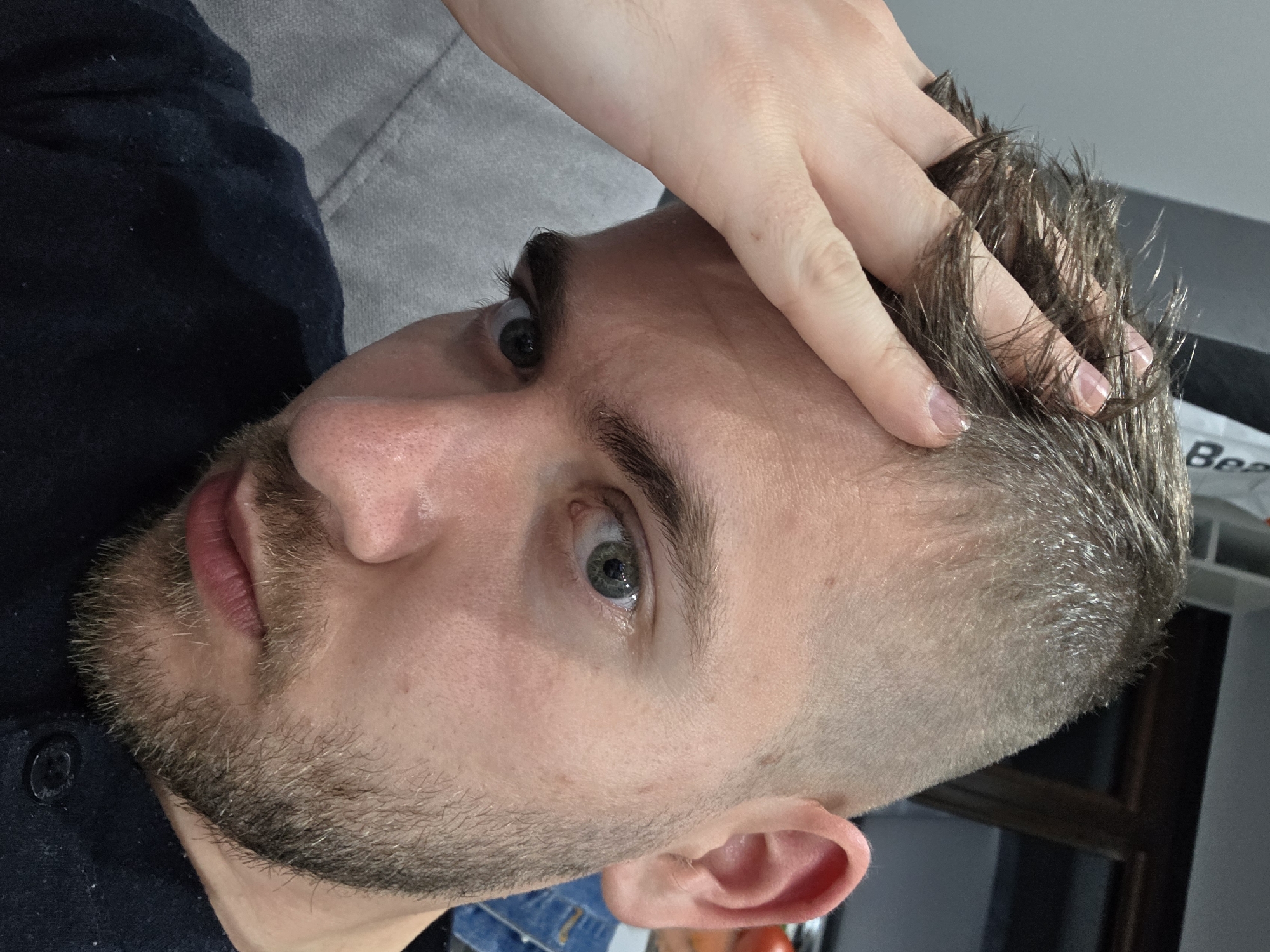 A close-up, tilted shot features a man with light skin, gray-green eyes, and short, dark brown hair. His hair is short on the sides, transitioning to longer strands on top, with a hand running through the top section. He has a beard and mustache with a mix of light and dark hairs. The man is wearing a dark-colored shirt, and a portion of what appears to be a denim jacket is visible in the bottom right corner. The background is blurred, showing parts of a gray couch and a glimpse of a white box with black lettering. The location is not ascertainable from the context.