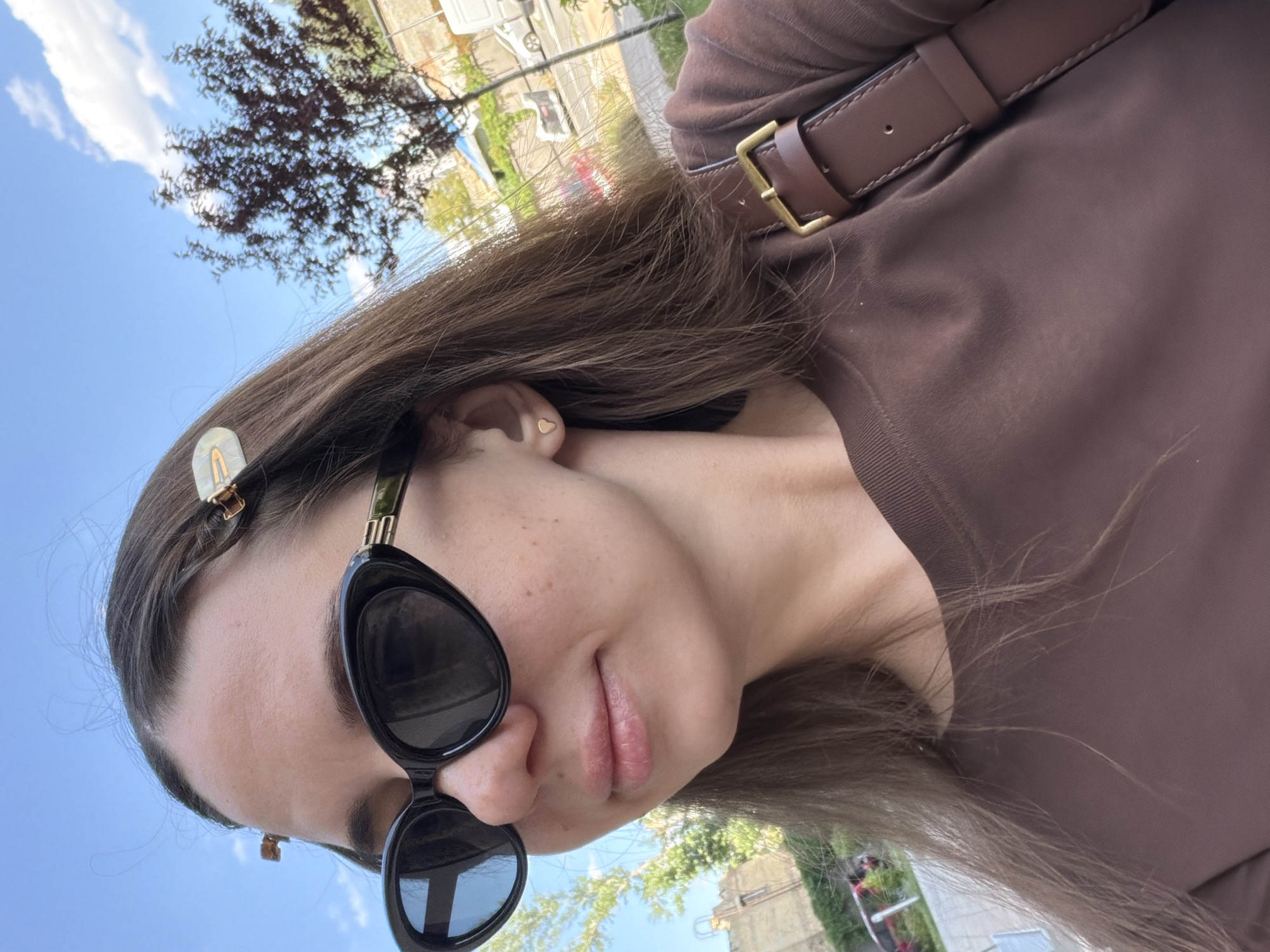A close-up, front-facing, outdoor selfie shows a woman with long, brown hair, wearing black, cat-eye sunglasses and a brown crew-neck t-shirt. Her hair is partly clipped back with small, decorative clips. She wears a small, heart-shaped earring in her visible left ear. A brown leather strap with a brass buckle, likely part of a bag or backpack, is visible across her right shoulder. The background suggests an urban environment with a clear blue sky, white clouds, trees, buildings, and a street with vehicles. The overall impression is that the photo was taken on a sunny day in a city, possibly Kyiv, Ukraine, given the context provided.