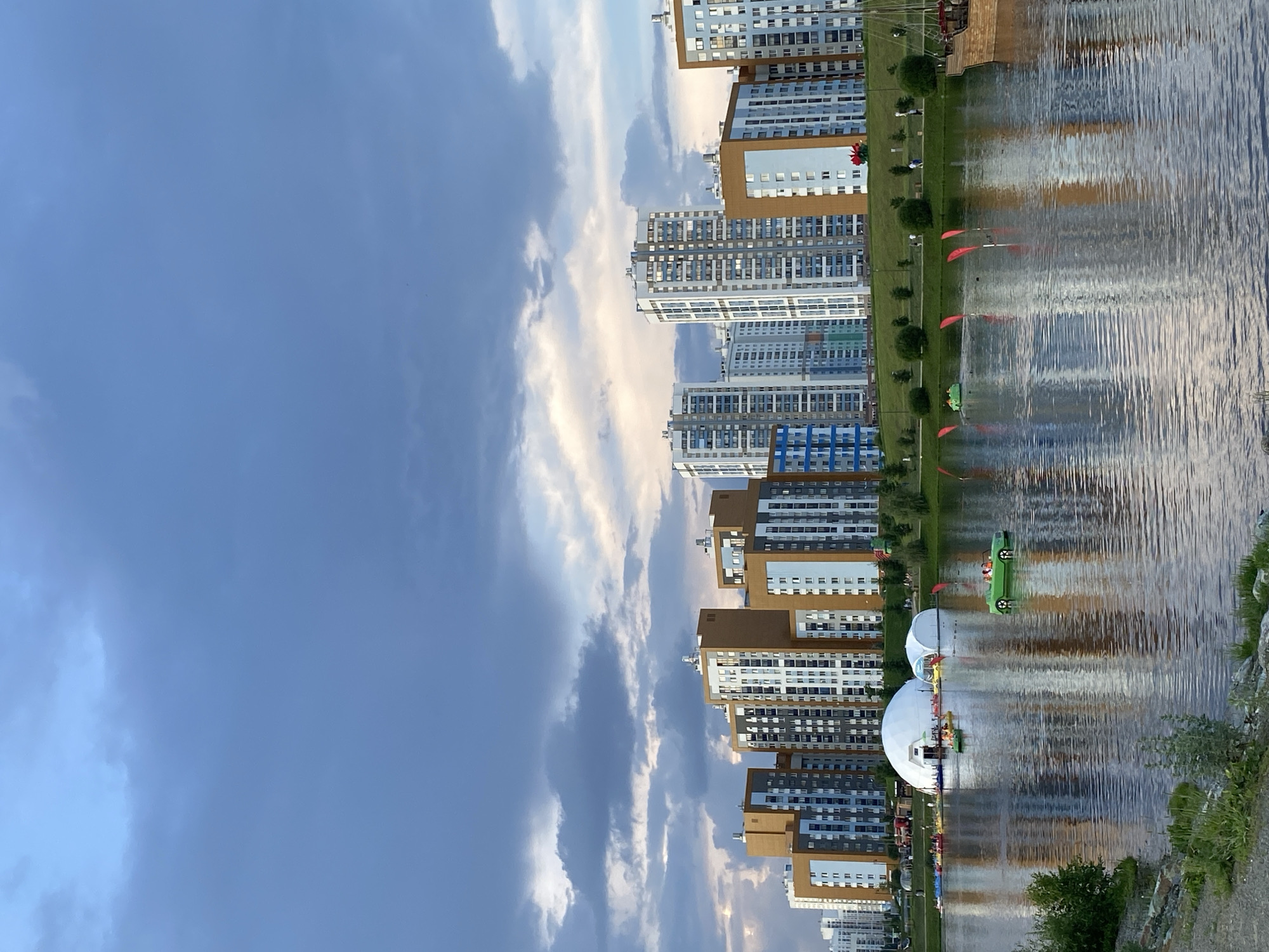 This is a photograph of the city of Yekaterinburg, Russia, showcasing a modern residential area situated along a body of water. The scene captures a twilight sky with dramatic clouds, reflecting on the calm surface of a lake. In the foreground, the water is dotted with several small pedal boats, including one shaped like a green convertible with several occupants. The shoreline is lined with green grass and trees. Dominating the background is a cluster of tall, modern apartment buildings with a mix of brown, white, and blue facades. Two large white dome-shaped structures, possibly temporary event venues, are visible on the lake's edge. Further to the right, a wooden ship with sails is docked. The overall impression is a serene urban landscape with recreational activities taking place on the water.