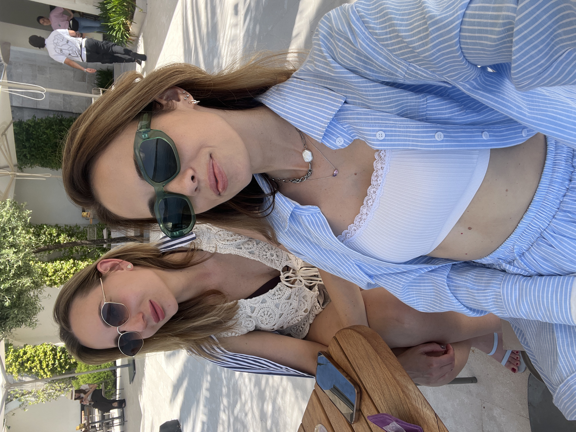 This media file presents a close-up, selfie-style image of two young women in an outdoor, sunny setting, likely a cafe or resort in Tivat, Montenegro. The image is rotated 90 degrees clockwise, giving the impression that the subjects are leaning into the frame.
The woman in the foreground has long, light brown hair and is wearing dark green, square-framed sunglasses. She is dressed in a light blue and white striped button-down shirt, worn open over a white ribbed crop top with delicate lace trim along the neckline. Her accessories include multiple ear piercings (studs and cuffs) and two layered necklaces: a chunky silver chain with a baroque pearl-like pendant, and a finer chain with a small, light pink or purple stone. She looks towards the camera with a relaxed expression and slightly parted lips.
Slightly behind and to her right is the second woman, with long, blonde-brown hair and round, gold-rimmed sunglasses. She wears a light-colored, intricate crocheted or lacy top over a darker undergarment. Her expression is similarly relaxed.
The background shows hints of a clean, modern outdoor environment with white architectural elements, lush green foliage, and other indistinct people. A wooden table occupies the lower right foreground, on which a metallic object and a purple item are visible. The lighting is bright and sunny, indicating daytime. The first woman's light blue sandals and red-painted toenails are also visible near the bottom right. No discernible text is present in the image. The overall atmosphere is relaxed and casual.
