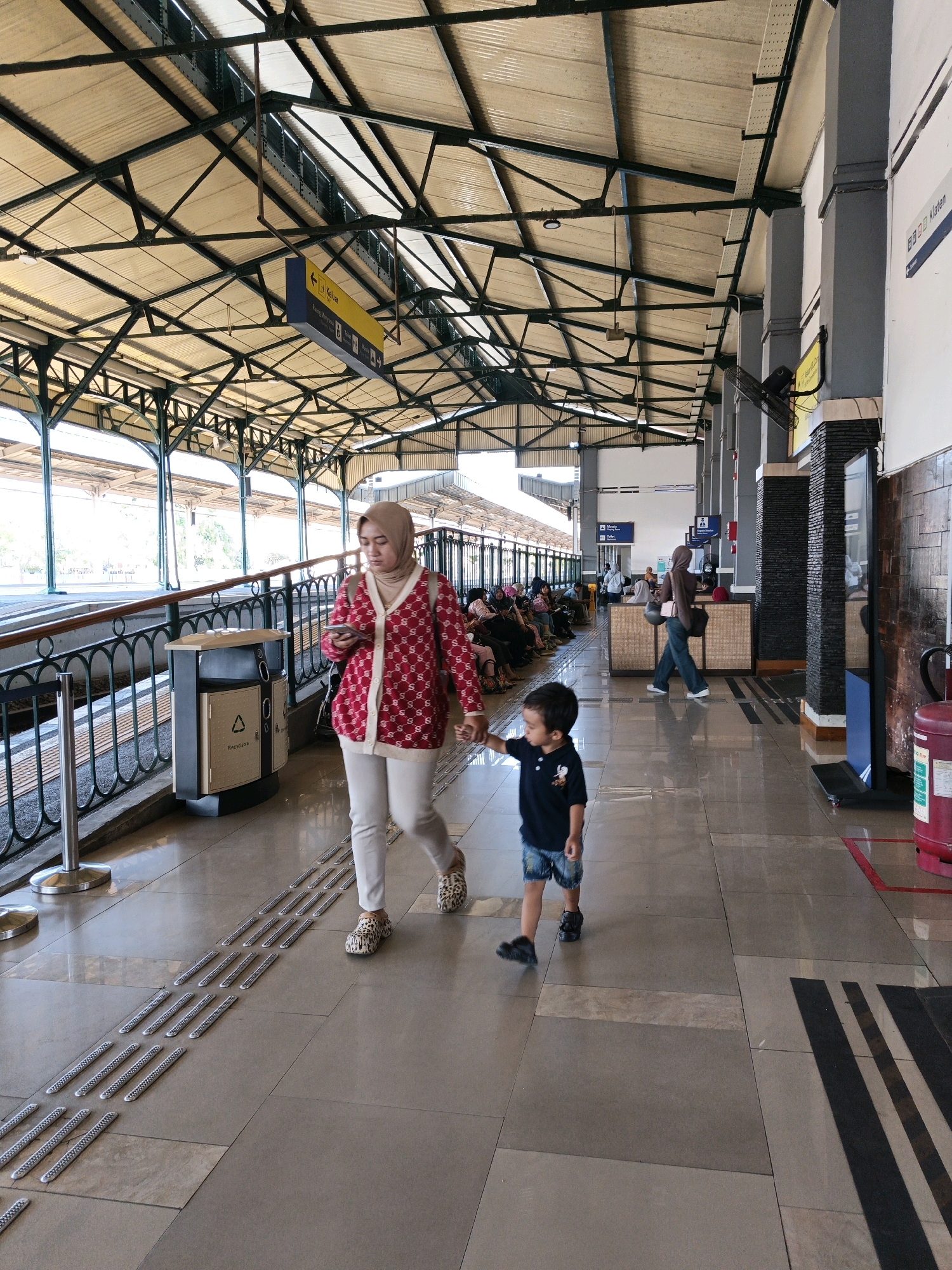 This image captures a bustling scene at the Klaten train station in Klaten, Indonesia. The foreground is dominated by a woman in a light-colored hijab and a red cardigan adorned with a repeating white pattern, who is holding a smartphone and walking alongside a young boy in a dark blue t-shirt and denim shorts. They are walking on a tiled platform that features tactile paving for visually impaired individuals.
In the background, other passengers are visible, some seated on benches and others standing. The train station's architecture features a prominent roof supported by metal beams and columns, with a yellow directional sign hanging overhead. Various objects, including trash bins and fire extinguishers, are placed along the platform. The overall environment suggests a typical day at a busy Indonesian train station.