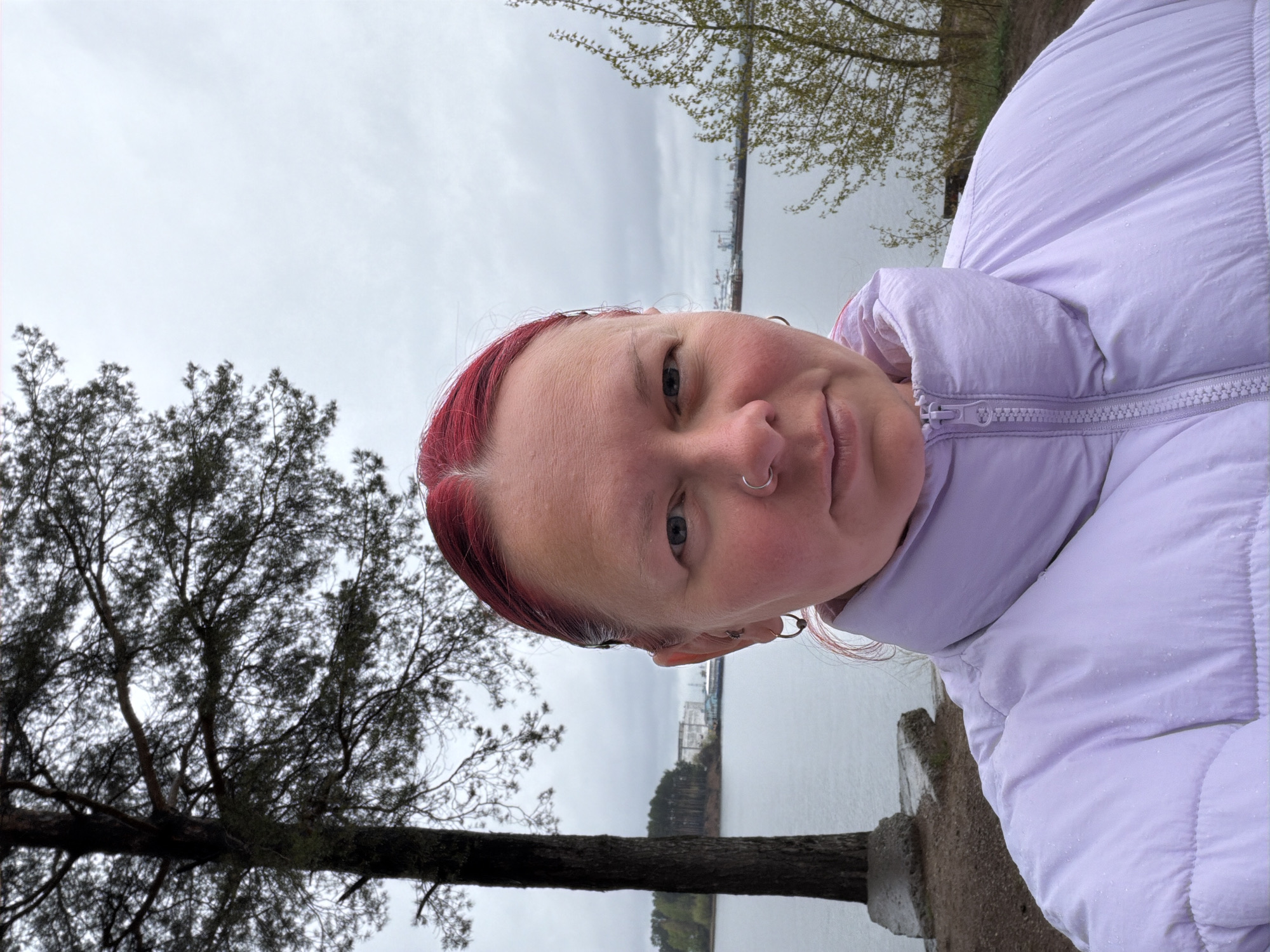 A medium shot shows a person with red hair and blue eyes wearing a light purple puffer jacket. The person's face is angled towards the camera, featuring a silver nose ring. The background reveals a body of water with land visible across it, under an overcast sky. A tree with bare branches is present on the left side of the frame. The location is Chaikovsky, Russia.