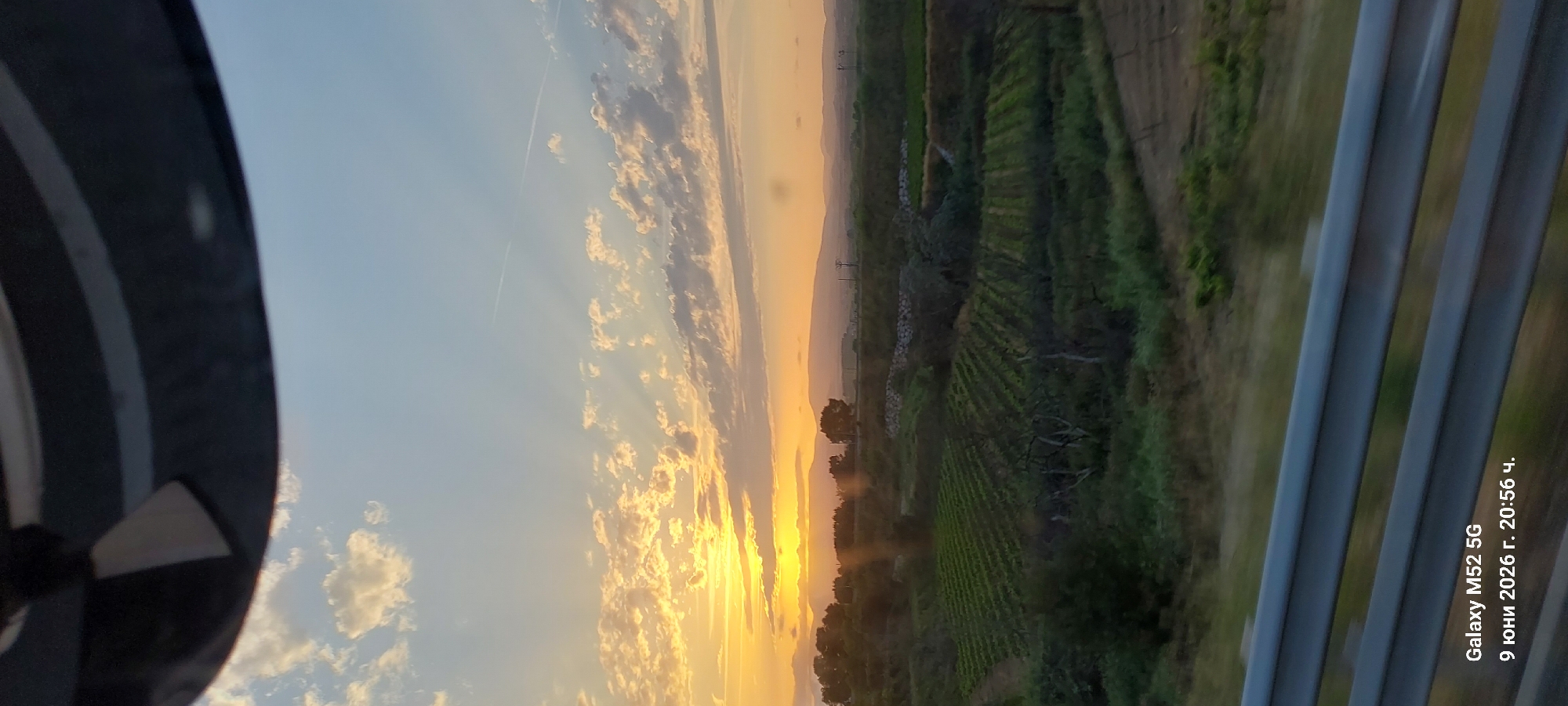 This is a photograph of a rural landscape at sunset, likely taken from a moving vehicle. The foreground features a metal guardrail. Beyond the guardrail, there are cultivated fields, possibly vineyards, with distinct rows of green plants. In the mid-ground, trees and rolling hills are visible. The background shows distant mountains. The sky is filled with dramatic clouds illuminated by the setting sun, casting warm orange and yellow light. Sunbeams are visible radiating from behind the clouds. The timestamp in the lower left corner indicates the photo was taken on June 9, 2026, at 8:56 PM and was captured by a Galaxy M52 5G. The location is identified as La Granada, Spain.