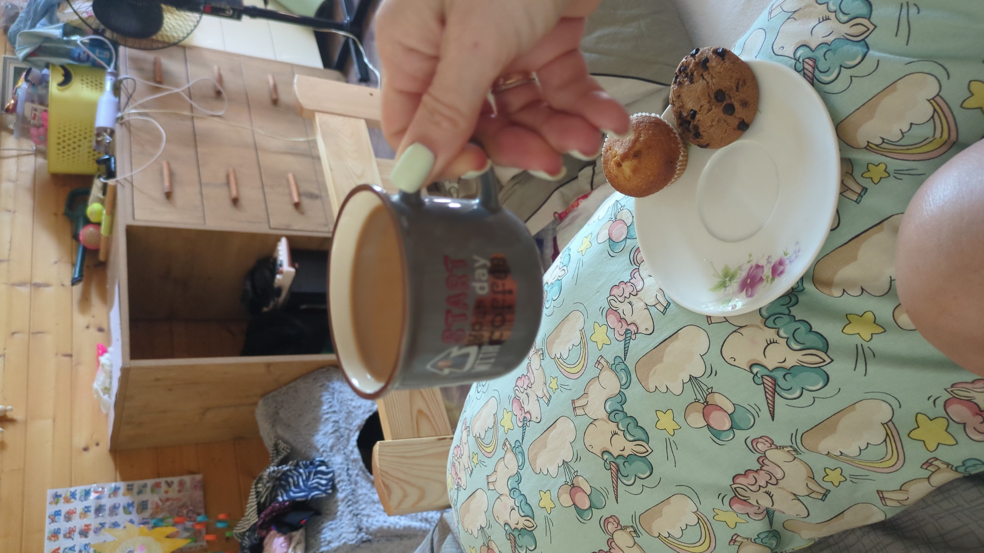 The image depicts an indoor scene, likely a bedroom or living area, with wooden paneled walls and a wooden dresser. A hand with light green nail polish is holding a dark mug that says "START YOUR DAY WITH COFFEE." A small white saucer with a floral design holds two baked goods: a plain muffin and a chocolate chip cookie. The lower part of the image shows a person's leg covered in light green fabric with a pattern of unicorns, rainbows, and stars. The background contains a bed with striped bedding, a gray pet bed, and a partially visible fan. The scene is casual and appears to be a moment of relaxation. There is no visible indication of the specific location being in Chișinău, Moldova.