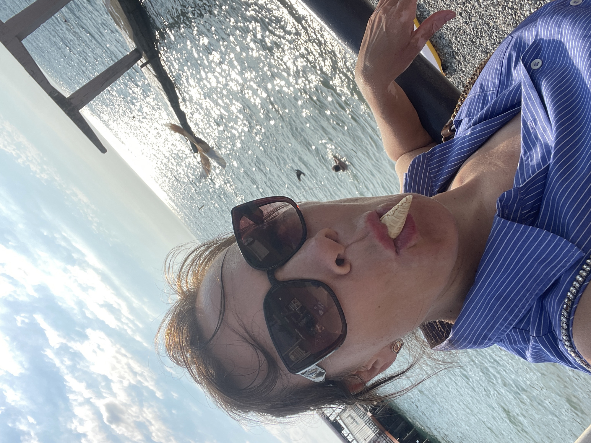 In the city of Sochi, Russia, on a sunny day, a woman with sunglasses and a blue striped shirt is taking a selfie by the sea. She has a piece of ice cream cone in her mouth. The sea is sparkling under the bright sun. There are several birds flying over the water, and a person can be seen swimming in the distance. There is also a wooden pier in the background. The woman is leaning on a railing, and the ground is covered with pebbles.