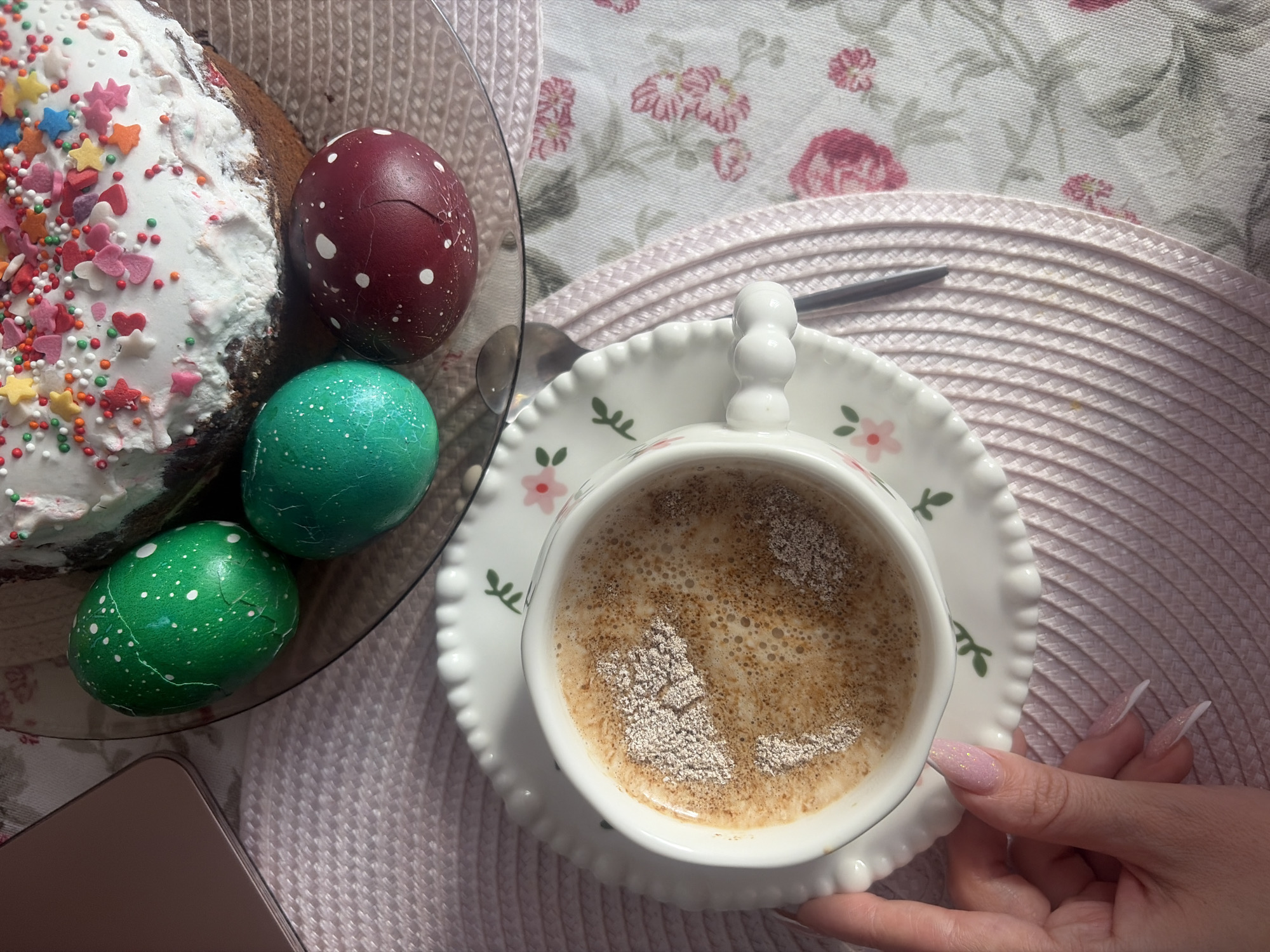 A high-angle shot captures a breakfast or brunch scene, potentially associated with Easter celebrations. The composition includes a slice of Easter bread cake decorated with white icing and colorful sprinkles, alongside a set of colored Easter eggs in green and maroon on a clear plate. Next to the plate is a cup of coffee with milk, held by a hand featuring long, manicured nails. The objects are arranged on a table with a floral patterned tablecloth and a woven placemat. The environment suggests a home setting, potentially located in Saint Petersburg, Russia, given the Eastern European Easter traditions.