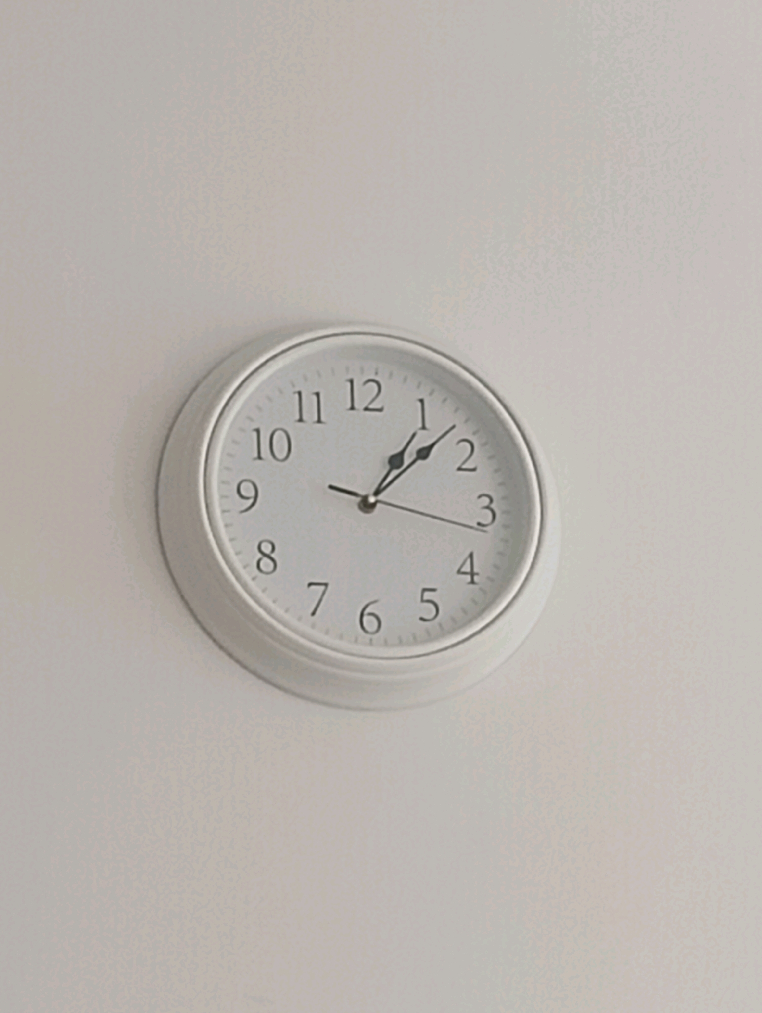 A white analog clock is mounted on a plain, off-white wall. The clock has black Arabic numerals indicating the hours from 1 to 12. The clock hands are black and are positioned at approximately 1:08. The image does not contain any identifiable landmarks or features that would place it within the city of Szczecin, Poland.