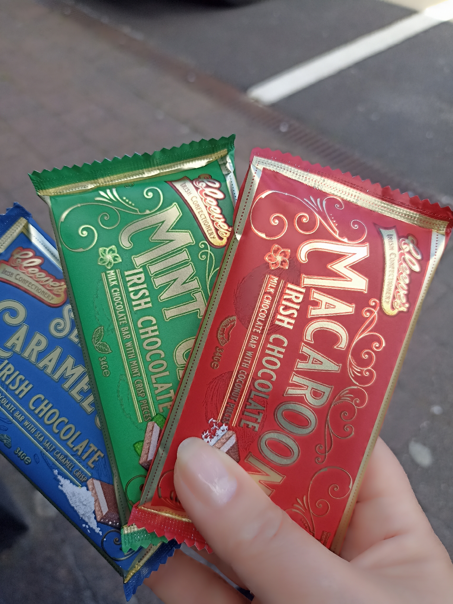 In Annagry, Ireland, a hand holds three chocolate bars. The bars, from left to right, are blue, green, and red. The blue bar is labeled "SEA CARAMEL IRISH CHOCOLATE BAR WITH SEA SALT CARAMEL CRISP". The green bar is labeled "MINT IRISH CHOCOLATE BAR WITH MINT CRISP PIECES". The red bar is labeled "MACAROON IRISH CHOCOLATE BAR WITH COCONUT PIECES". All three bars feature gold lettering and decorative patterns. The background shows a blurred grey surface, possibly a street or pavement.