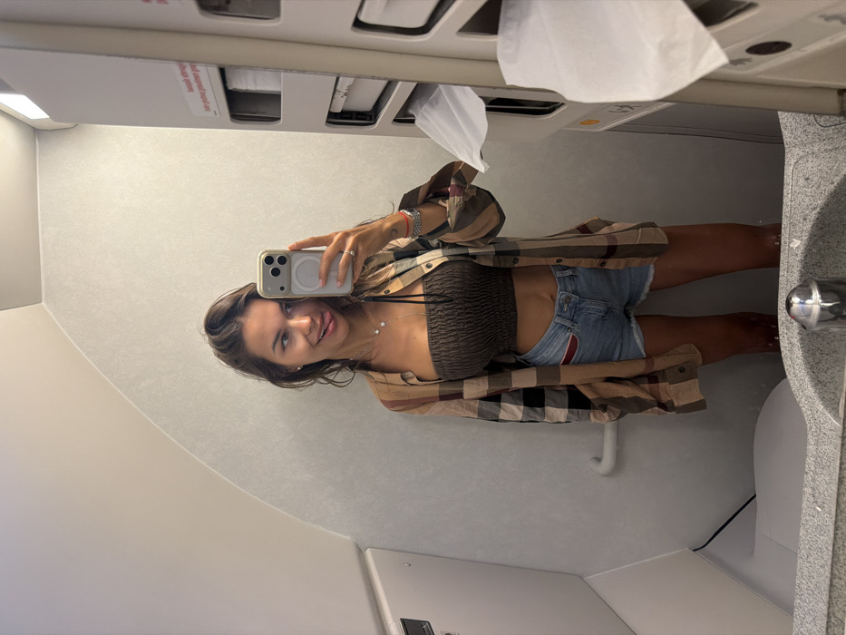 A woman in a plaid shirt, brown tube top, and denim shorts poses in an airplane lavatory mirror selfie. She's holding a phone with a visible MagSafe charger. The lavatory features a sink, faucet, toilet, and overhead compartment. A partially dispensed paper towel hangs from the dispenser above. The lighting is bright, typical of an airplane lavatory. Her expression is playful.