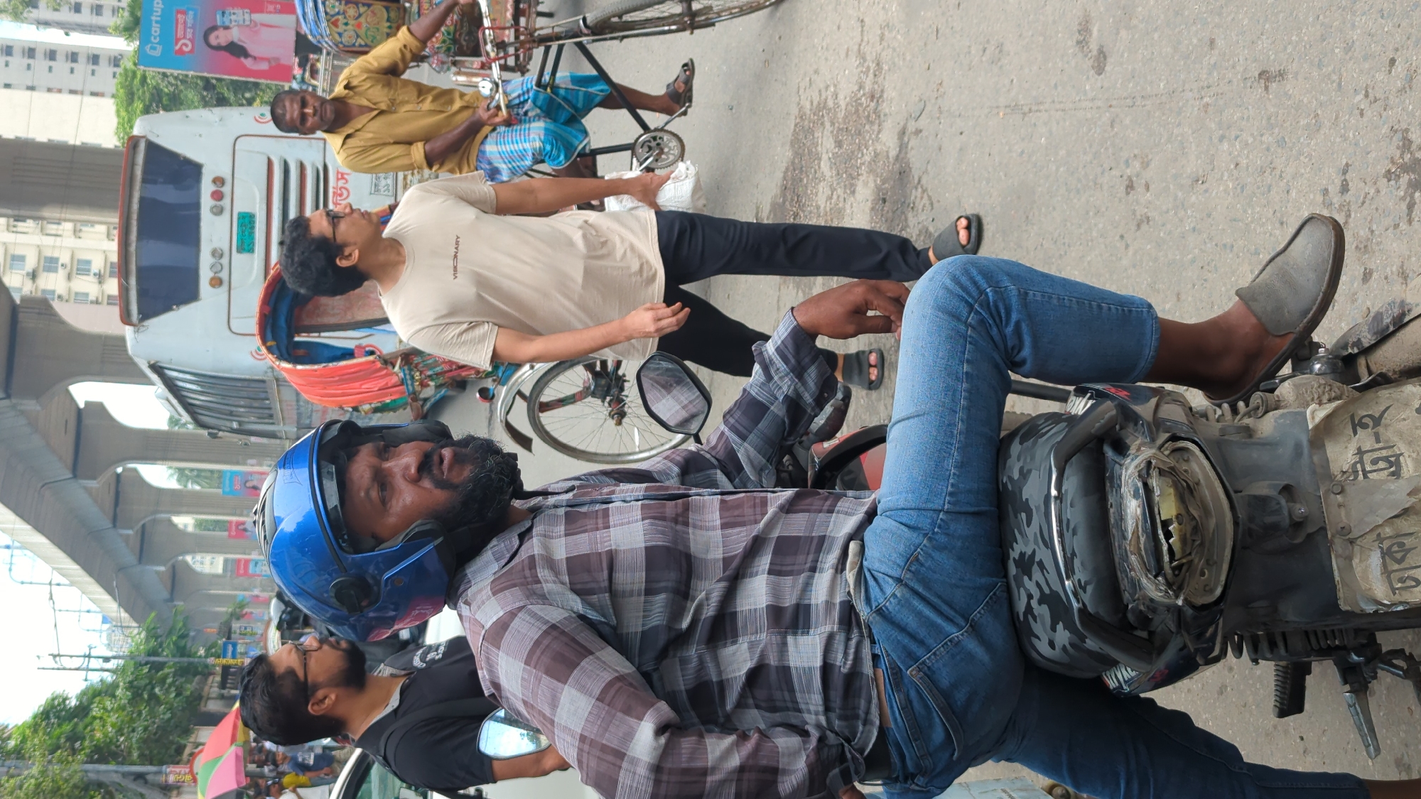 In Dhaka, Bangladesh, a man wearing a blue helmet and a checkered shirt sits on a motorcycle. He has a beard and is looking to the right. He is wearing blue jeans and grey slip-on shoes. In the background, a bus and several rickshaws are visible, along with an elevated road structure. Other people are walking and standing in the scene. The environment appears to be a busy street in an urban area.