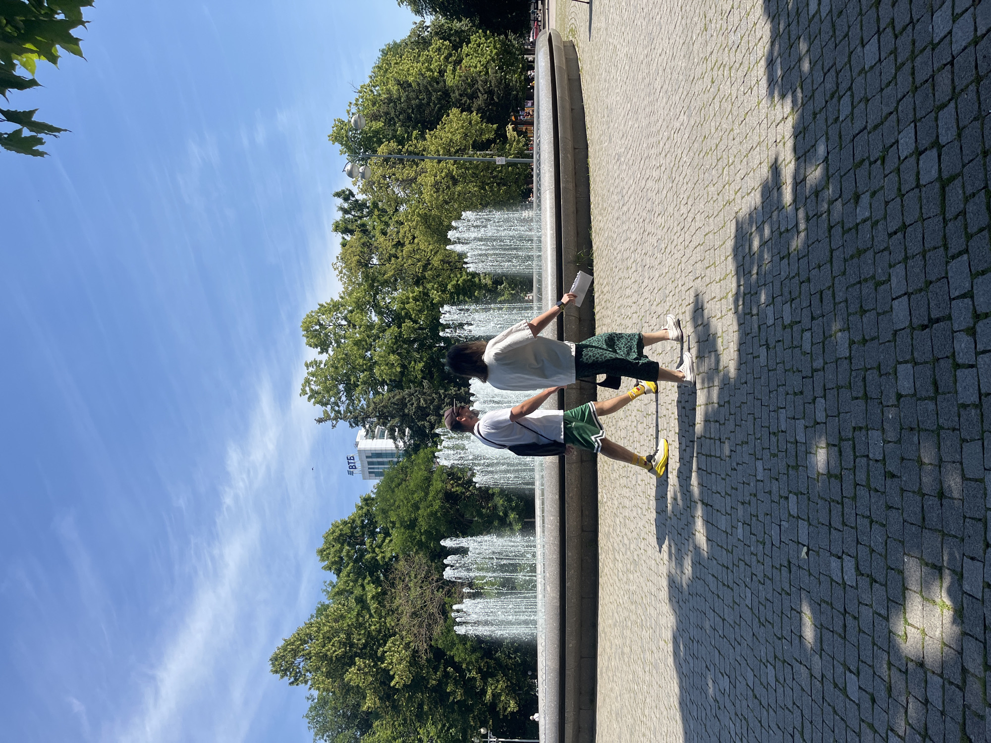 Two individuals walk across a cobblestone plaza in front of a large, circular fountain. The fountain features multiple streams of water arching upwards. Behind the fountain, lush green trees form a dense backdrop, with a modern white building bearing the Cyrillic lettering "ВТБ" (VTB) visible above the trees in the distance. The sky is blue with scattered white clouds. The plaza is paved with dark grey cobblestones, and the foreground is partially shaded by dappled sunlight filtering through unseen foliage. The man on the left wears a white t-shirt, green shorts with white trim, yellow socks, and bright yellow athletic shoes. He carries a black backpack. The woman on the right wears a white shirt and a green patterned skirt. She holds a white object, possibly papers, in her right hand. The overall scene suggests a pleasant day in an urban park setting. The VTB building suggests a location in Russia, and the architectural style and climate are consistent with Krasnodar.