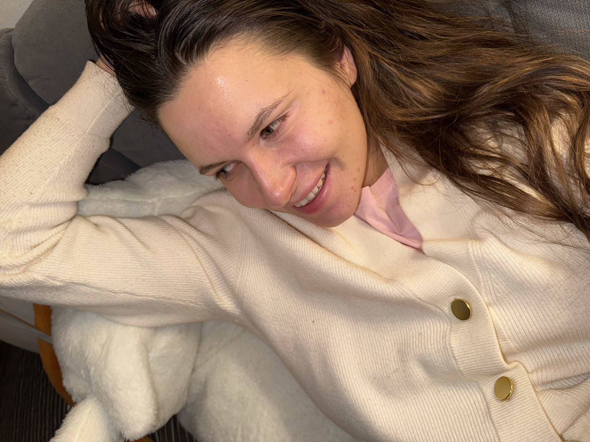 A young woman lies on a plush white surface wearing a light yellow cardigan over a pink top. She has long brown hair and is smiling. She appears to be indoors, resting on a sofa. The location is not discernible.