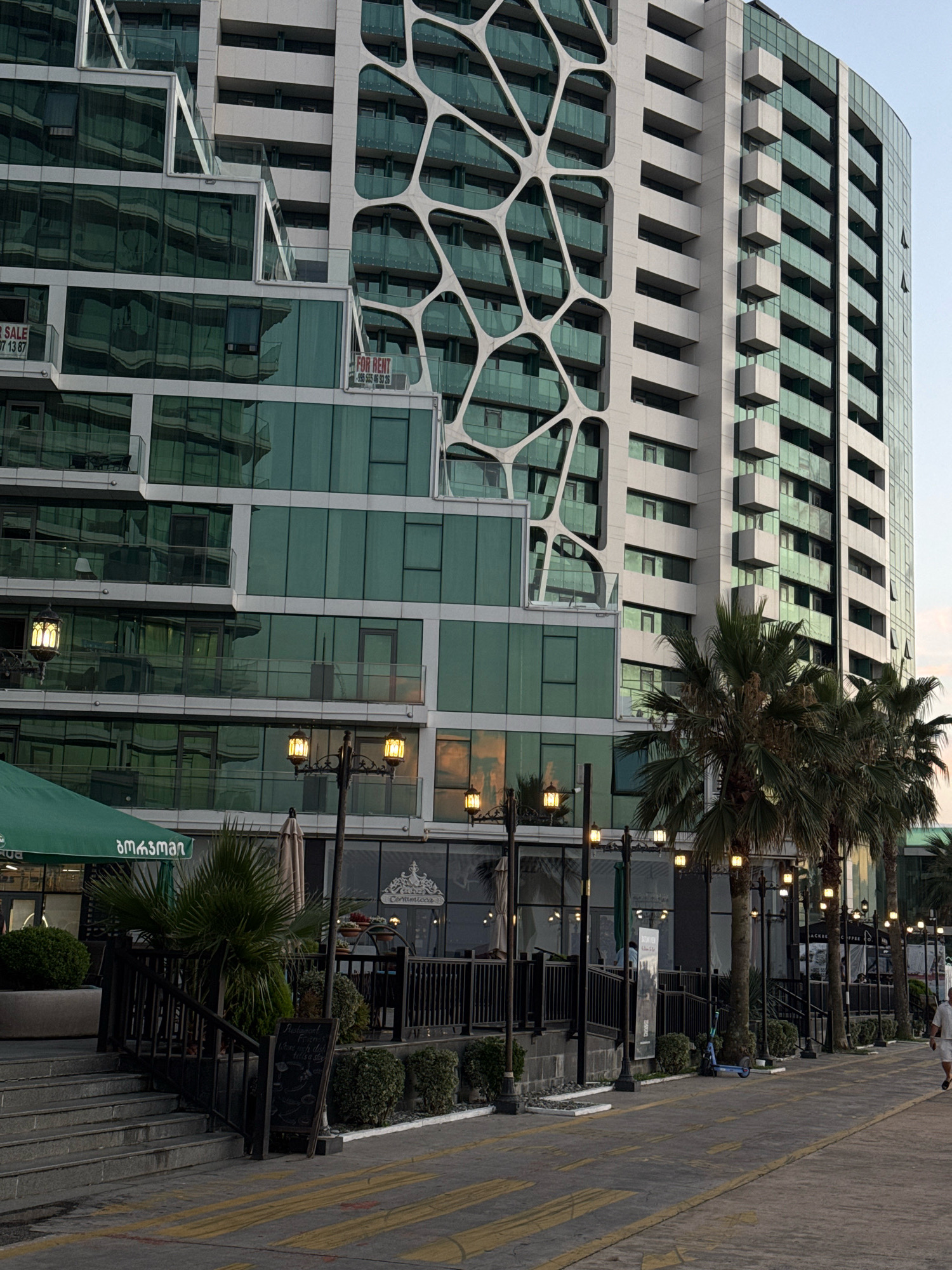 The image depicts a modern architectural building with a distinctive organic-shaped white facade overlaying a glass-covered structure. The building appears to be a residential or commercial complex, with multiple floors featuring balconies and large windows. The lower levels show storefronts and outdoor seating areas, suggesting commercial spaces. Palm trees line the walkway in front of the building, characteristic of a coastal or resort area.
The scene is set in Batumi, Georgia, a city known for its contemporary architecture and seaside promenade. The specific location is likely along the Batumi Boulevard, a popular pedestrian area that runs along the Black Sea coast. This area is characterized by its blend of modern buildings, green spaces, and public amenities.
In the foreground, a paved walkway with yellow markings is visible, indicating a pedestrian or cycling path. A person is walking away from the camera on the right side of the walkway. Several streetlights with decorative lanterns are positioned along the promenade. To the left, there are stairs leading up to an entrance, with a green awning extending from a building. A blue electric scooter is parked near the walkway.
The overall impression is one of a vibrant urban environment, likely during the late afternoon or early evening, given the lighting conditions and the sky. The architecture of the building stands out as a prominent feature, reflecting the city's modern development.