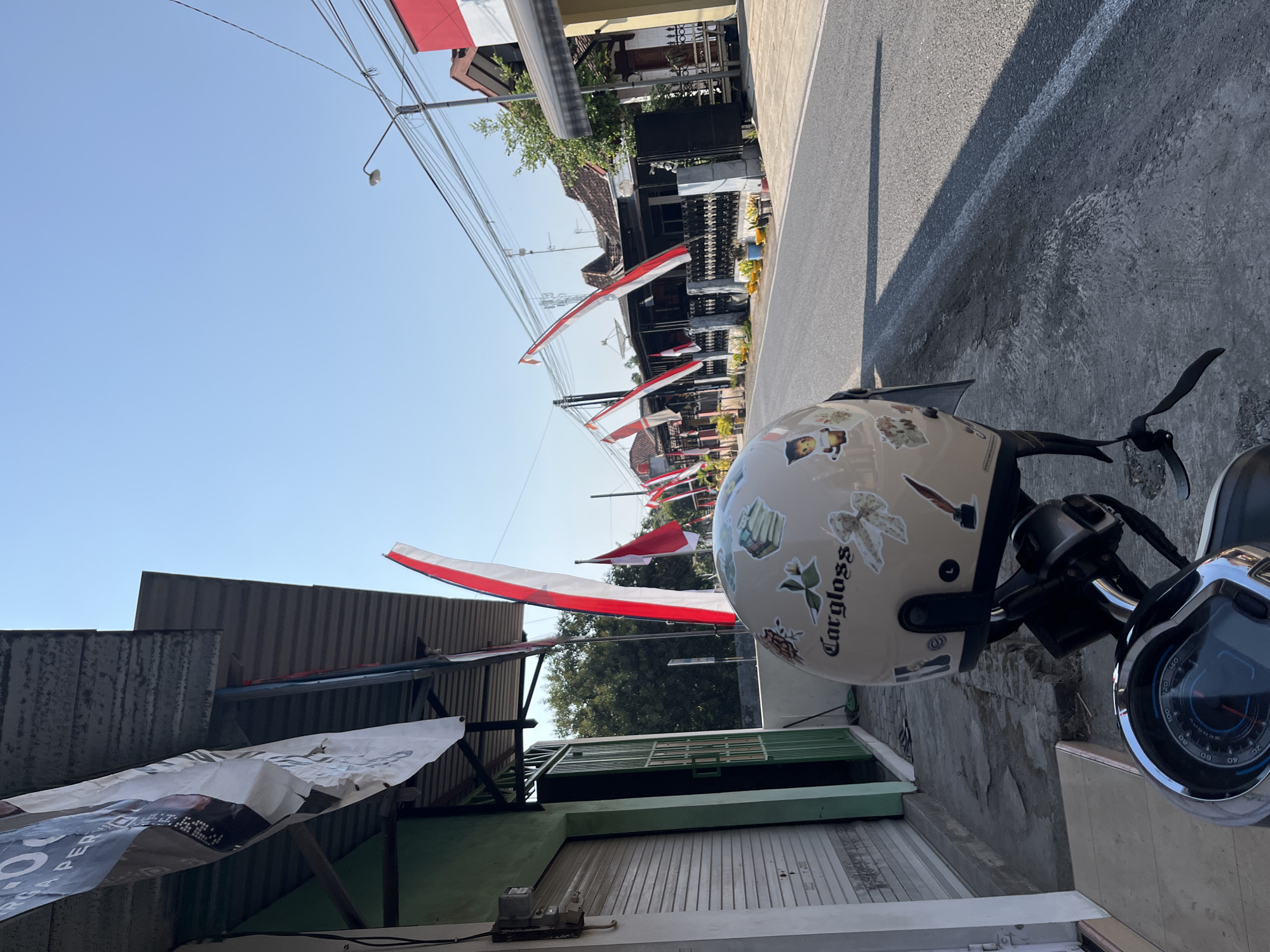 This is a full, eye-level shot of a street scene in Margorejo, Indonesia, under a clear blue sky. In the foreground, a scooter is parked on the sidewalk, its handlebars and dashboard visible. A beige helmet, adorned with several stickers, rests on the scooter's seat. The helmet has the word "Cargloss" written on it.
To the left of the scooter is the exterior of a building with a green door and a gray, corrugated metal shutter. A banner hangs from the roof of this building.
The street itself is paved and appears to be an asphalt road. Along the right side of the road, houses with dark roofs and some red and white decorations are visible. Numerous red and white flags are strung along poles lining the street, suggesting a festive or national celebration. The flags are the national flag of Indonesia. A street lamp and power lines are also visible in the background. The overall impression is a sunny day in a residential and commercial area of the city.