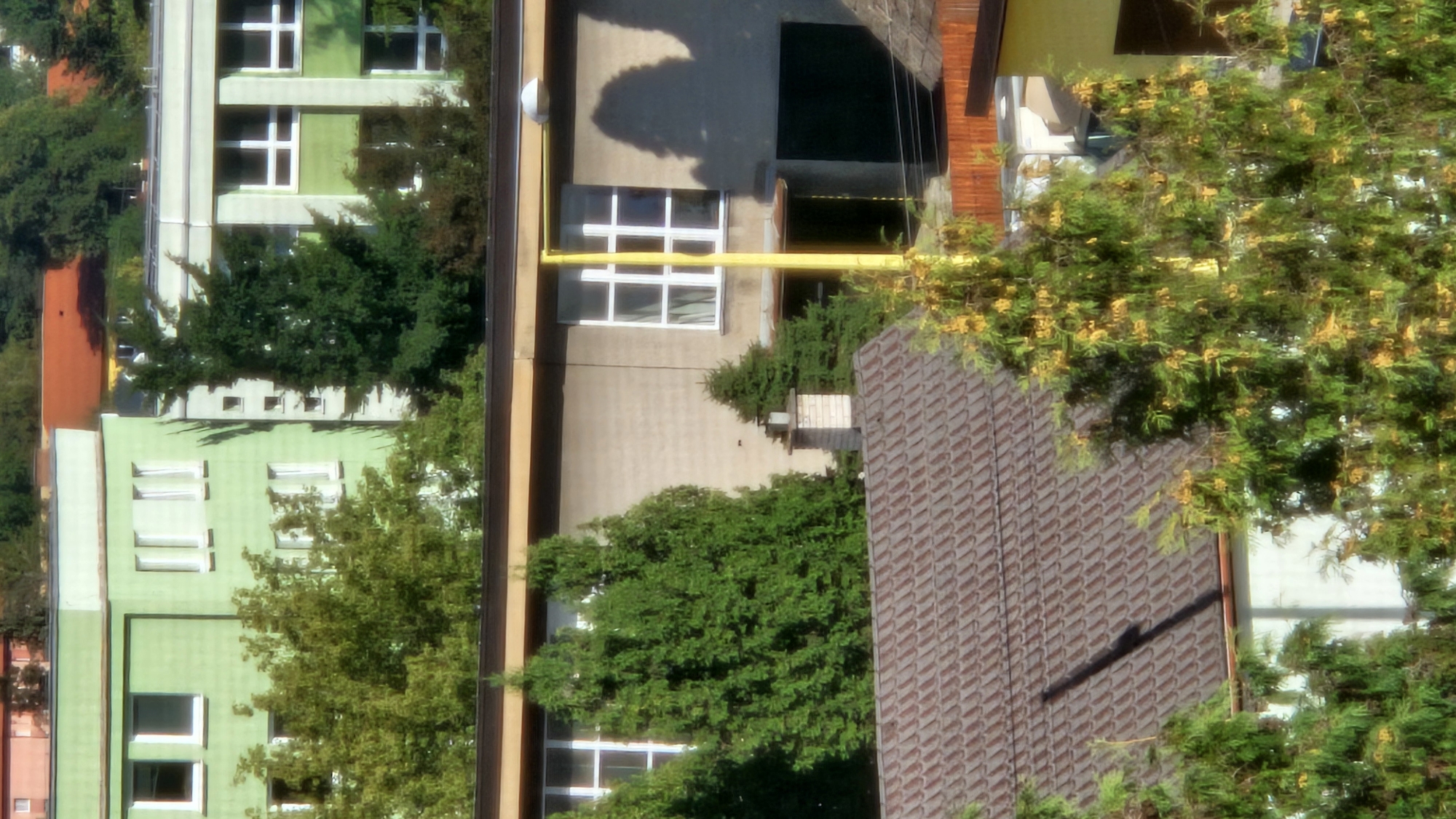 The image shows a multi-story building with a light green facade and white framed windows. There are trees in front of the building, some of which are tall and green. A section of a building with a terracotta tile roof is visible in the foreground. The overall scene suggests an urban or suburban environment, possibly a residential or educational complex given the building design and presence of trees. This specific location is in the municipality of Prešov, Slovakia.