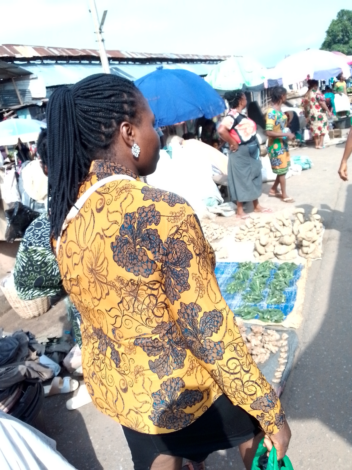 The image depicts a street market scene in Nkpor, Nigeria. A woman in the foreground, facing away from the camera, wears a yellow shirt with a floral pattern and black shorts. She has her hair braided into a ponytail and is wearing a large, ornate earring. Her right hand is holding a green plastic bag. Behind her, a woman in a patterned green and yellow dress stands, and further back, other individuals are visible under various market stalls with umbrellas and corrugated iron roofing. Piles of what appear to be yams and okra are displayed on the ground, some on blue tarpaulins. The ground is paved, suggesting a market area within the city.