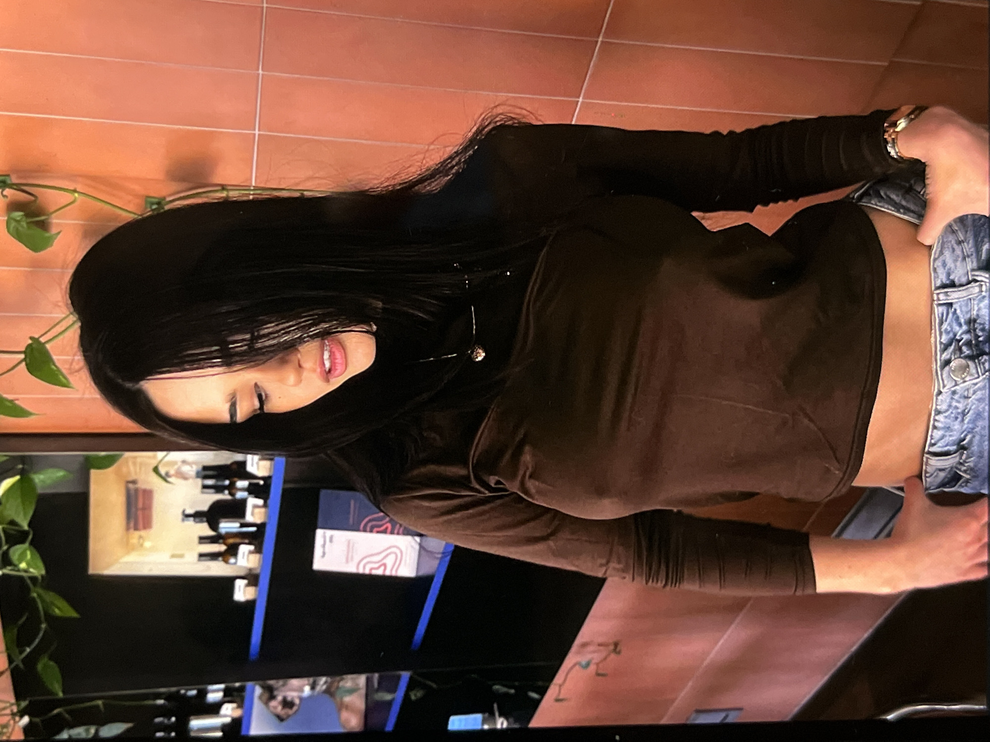 A young woman with long, dark hair stands in a dimly lit room. She is wearing a brown, long-sleeved, form-fitting shirt and blue jeans. Her head is tilted down, and her mouth is slightly open. She has a delicate necklace with a small pendant around her neck. To her left, shelves are visible, holding various bottles and some books or pamphlets. The wall behind her is tiled in a pale orange color. A plant with green leaves hangs from the top of the frame. The overall lighting suggests an indoor, possibly boutique or salon-like setting.