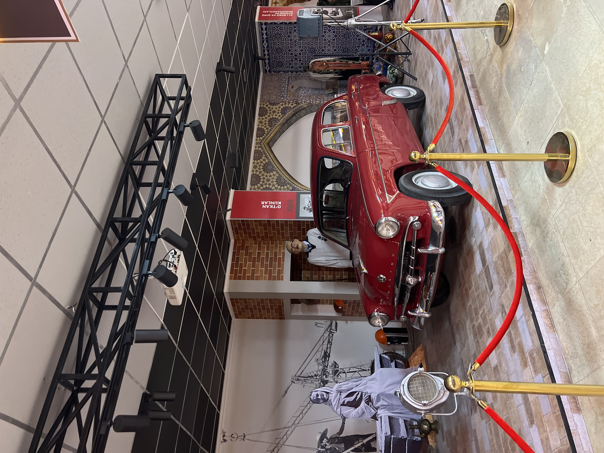 This image appears to be taken inside a museum exhibit in Tashkent, Uzbekistan, featuring vintage automobiles and historical displays. The main subject is a red vintage car, cordoned off by red velvet ropes attached to golden stanchions. Adjacent to the car, there is a mannequin dressed in period clothing, positioned inside a brick-lined alcove with a red sign above that says "O'TKAN KUNLAR." The background showcases large photographic prints depicting scenes related to the history of construction, including images of cranes. The museum's walls are tiled with a geometric pattern. The scene includes a complex lighting system supported by a black metal truss structure attached to the ceiling and a wall-mounted projector.