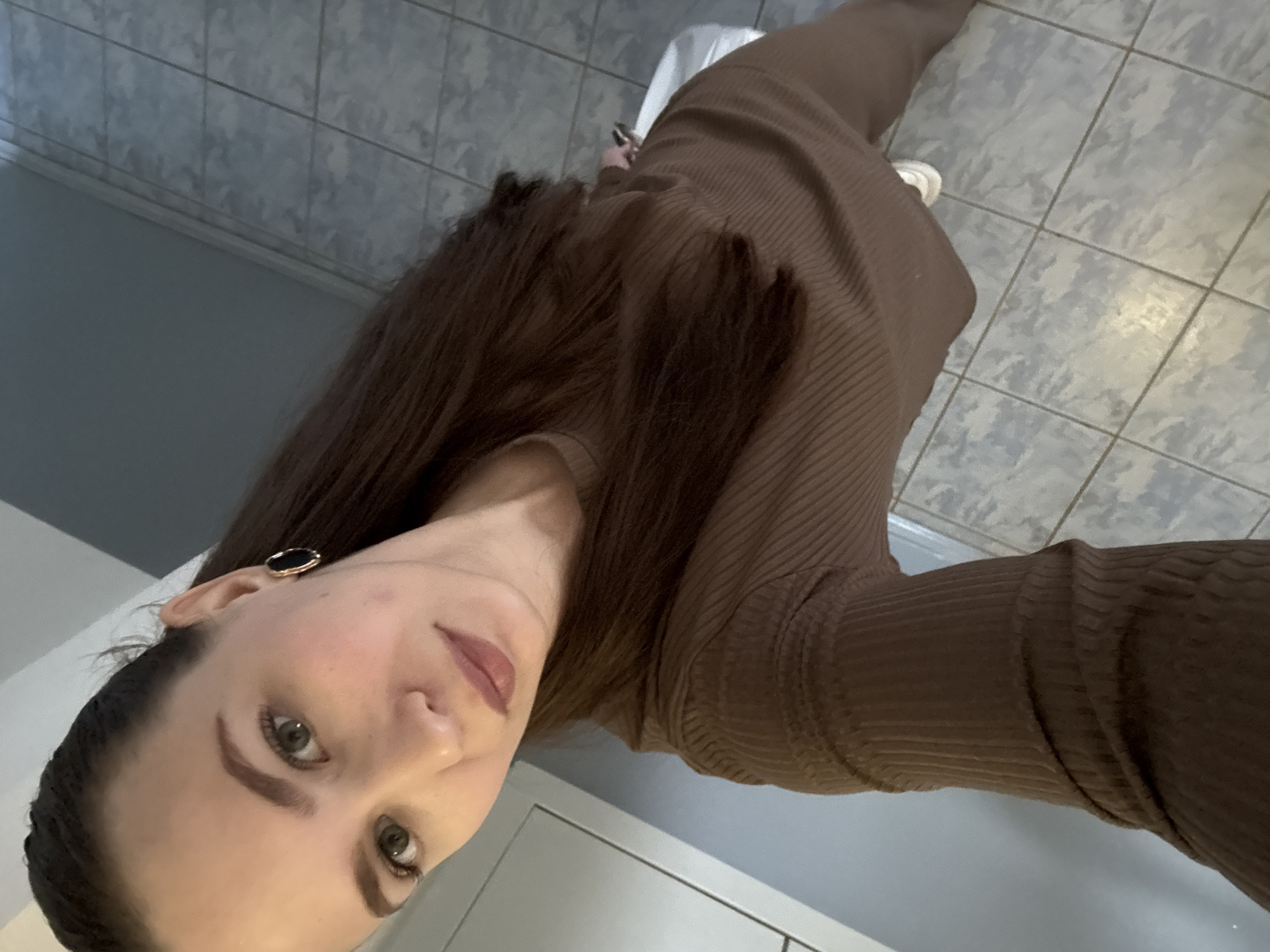 A young woman is captured in a close-up, full-body selfie taken indoors in Chaikovsky, Russia. The image is oriented sideways, with the woman appearing to be positioned vertically if corrected. She has long, straight, dark brown hair that cascades over her shoulders and down her back. Her head is tilted slightly, and she looks directly at the camera with a neutral expression. She has clear skin, pronounced eyebrows, and her lips are painted with a subtle pinkish-brown lipstick. A striking black and gold oval-shaped earring is visible on her left earlobe.
She is wearing a form-fitting, long-sleeved ribbed dress or a co-ord set in a rich dark brown or taupe color. The ribbed texture of the fabric is clearly visible across her torso and sleeves. Her left hand, possibly holding a phone, is partially visible near her hip in one of the cropped images. Her feet, clad in white sneakers, are also visible at the bottom of the frame, hinting at a casual ensemble.
The background consists of a light grey tiled floor with a subtle, aged pattern and dark grout lines. A plain, light grey wall borders the floor, suggesting an unadorned interior space, possibly a hallway or room within a residential building. The lighting is even, suggesting an indoor setting without harsh direct sunlight. No discernible text is visible in the image.
