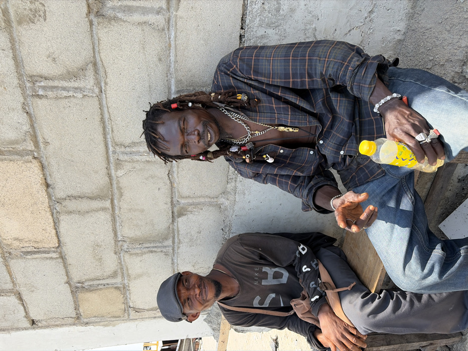 In Igboefon, Nigeria, two adult males are seated on a wooden bench outdoors, against an unpainted concrete block wall, under bright daylight.
The individual on the left is a dark-skinned male wearing a dark grey flat cap, a dark grey long-sleeved shirt with "NIKE SB" branding, and dark trousers. A brown shoulder bag strap is visible. He smiles, looking towards the individual on his right, with his hands clasped.
The individual on the right is a dark-skinned male with dreadlocks adorned with numerous colorful beads and charms. He wears an unbuttoned dark plaid long-sleeved shirt, revealing a dark undershirt, blue jeans, multiple silver-colored necklaces with a distinctive gold-colored pendant, several silver rings, and silver bracelets. He holds a clear plastic bottle with a yellow cap and yellow liquid in his left hand, and his right hand is slightly raised and open. He smiles, looking towards the camera.
The immediate background consists of a coarse concrete block wall with visible mortar lines. A light-colored vertical structural element is present on the far left.